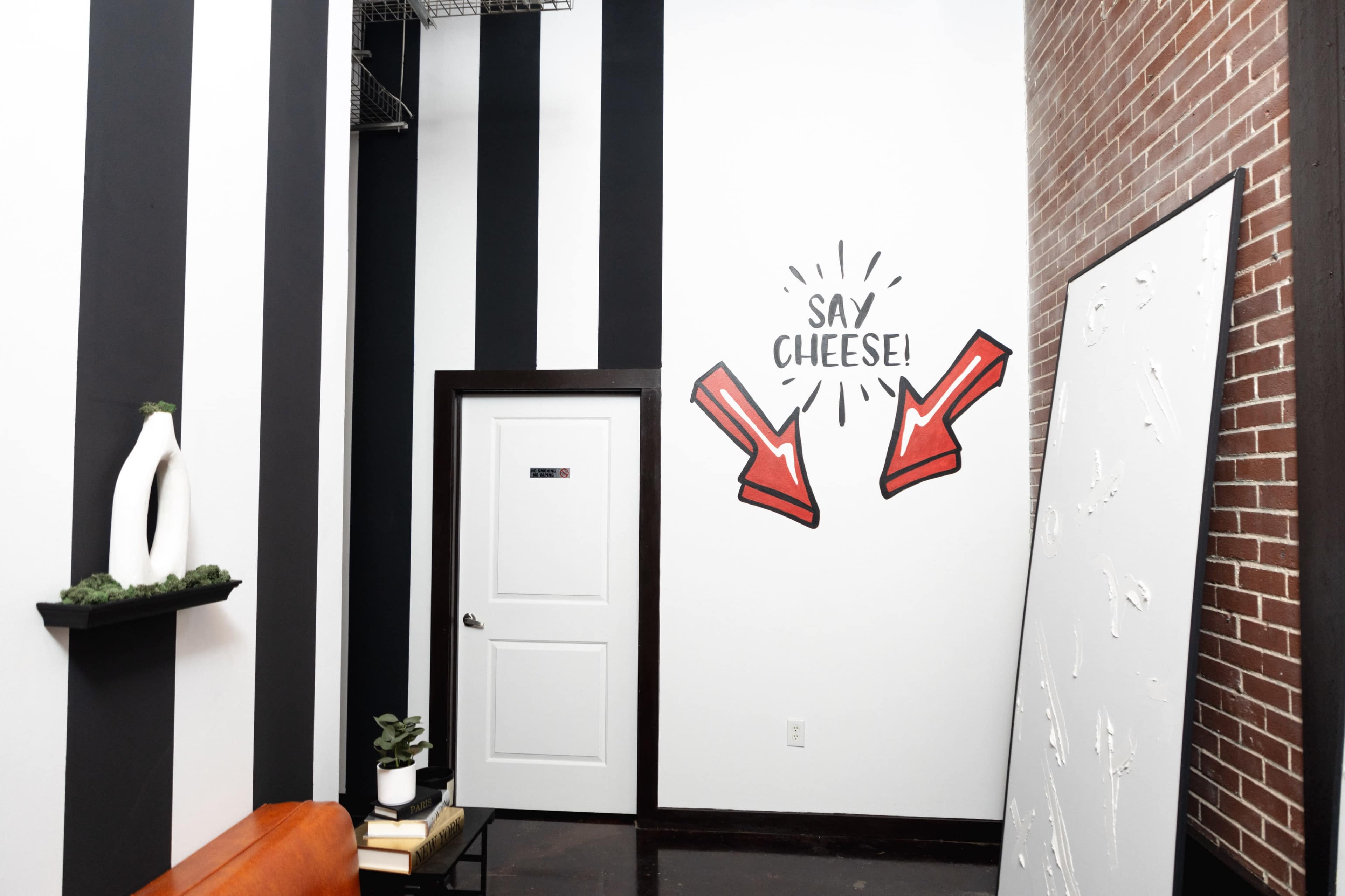 A room with vertical black and white striped walls, featuring a door with a sign and a painted graphic of two arrows pointing at the words "Say Cheese!" on one wall.