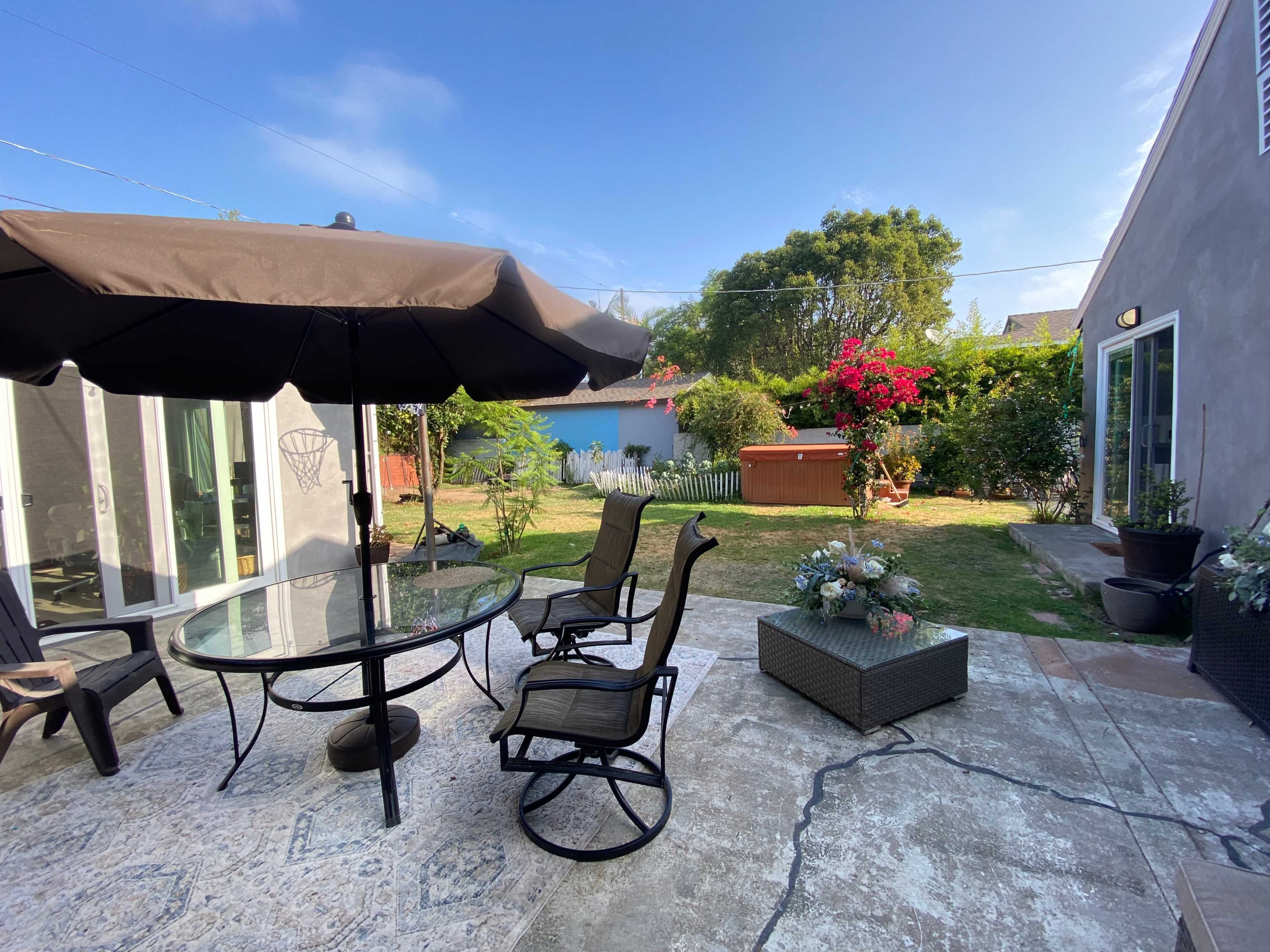 A patio area features a glass table with chairs under a large umbrella, surrounded by greenery and flowering plants.