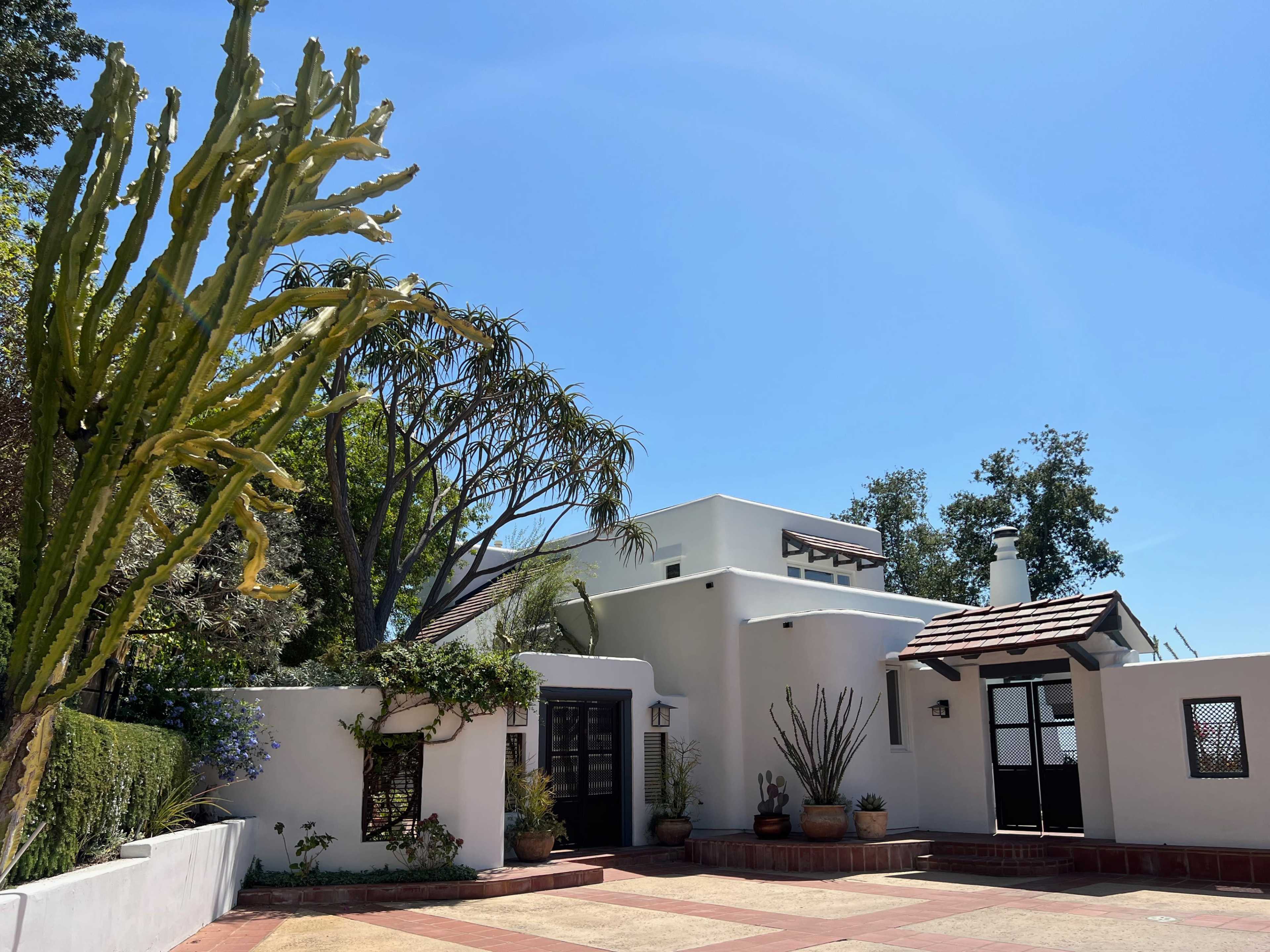 A white stucco house with a tiled roof is surrounded by various plants and trees under a clear blue sky.