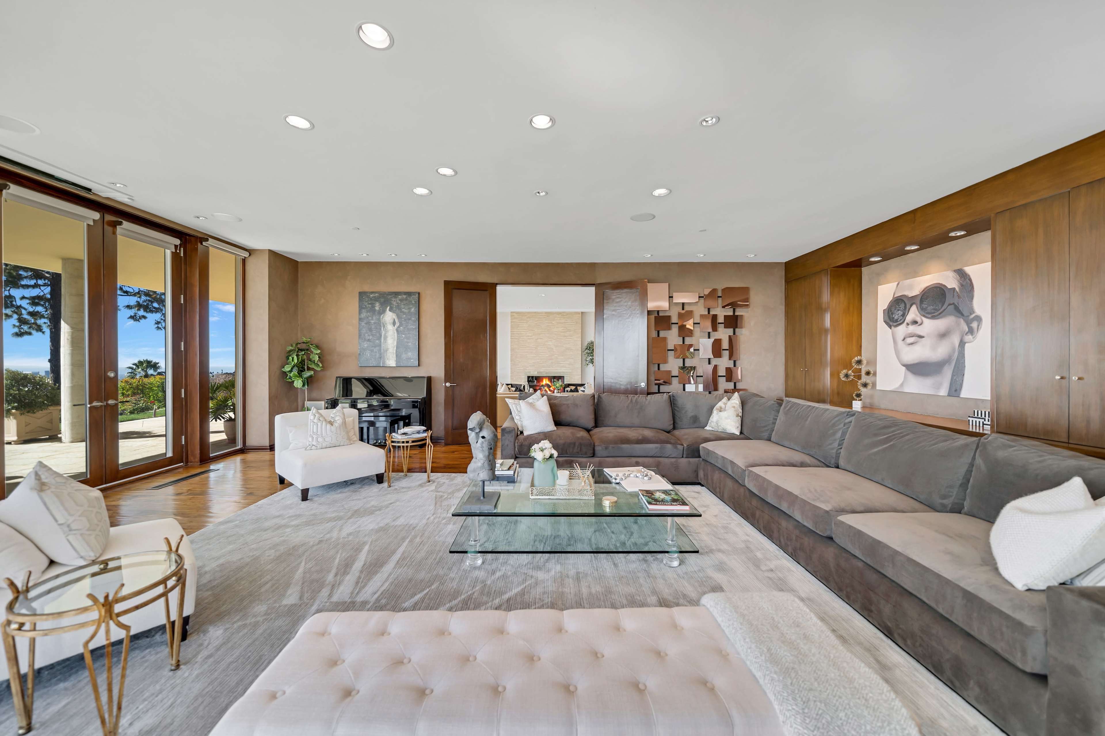 The spacious living room features a large sectional sofa, a glass coffee table, and a large artwork on the wall, with floor-to-ceiling windows that provide a view of the outdoors.