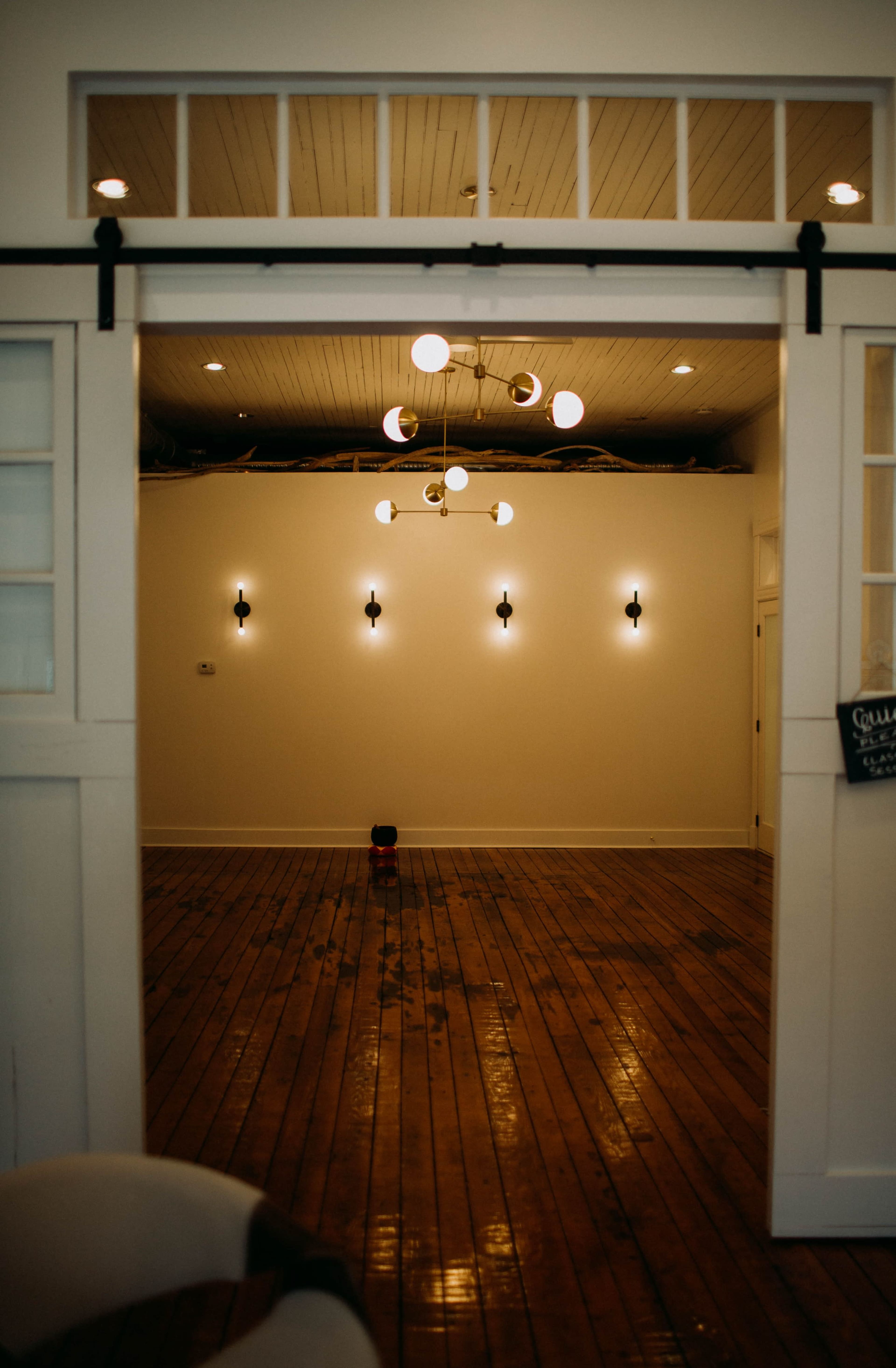 The image shows an empty room with wooden floors, a chandelier, and wall sconces illuminating the white walls.