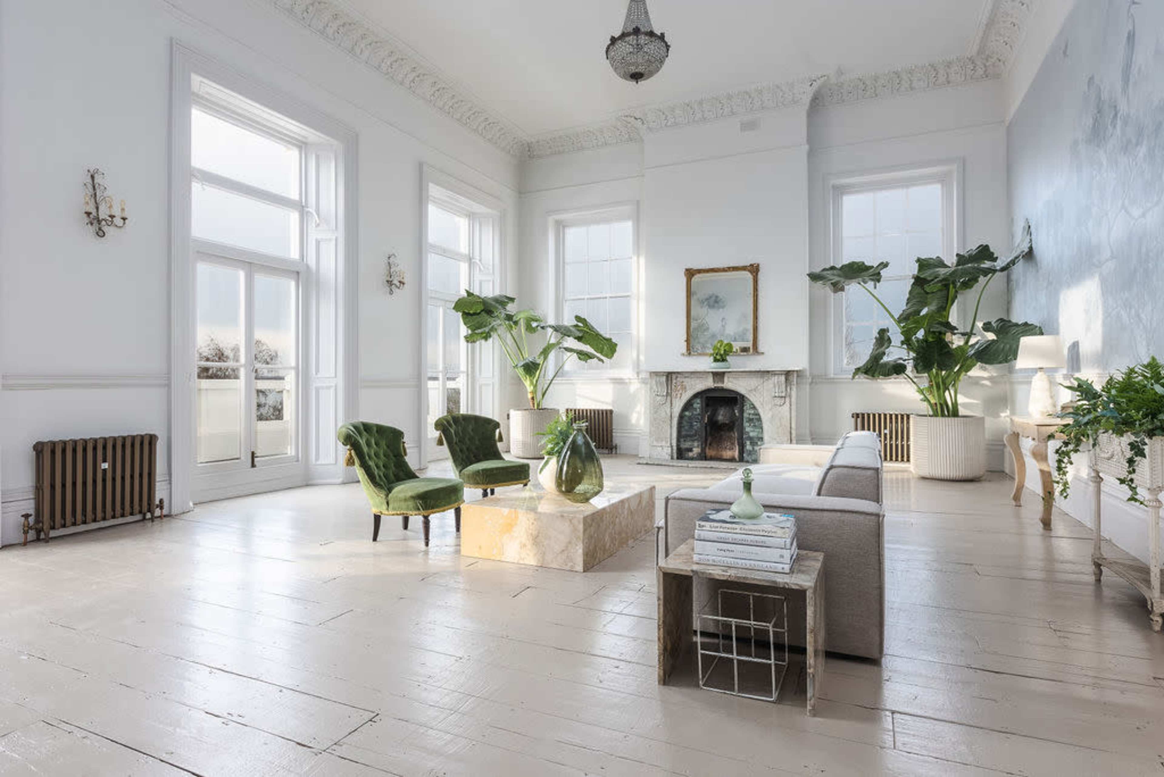 A spacious, brightly lit living room with large windows, two green chairs, a beige sofa, and a decorative fireplace.