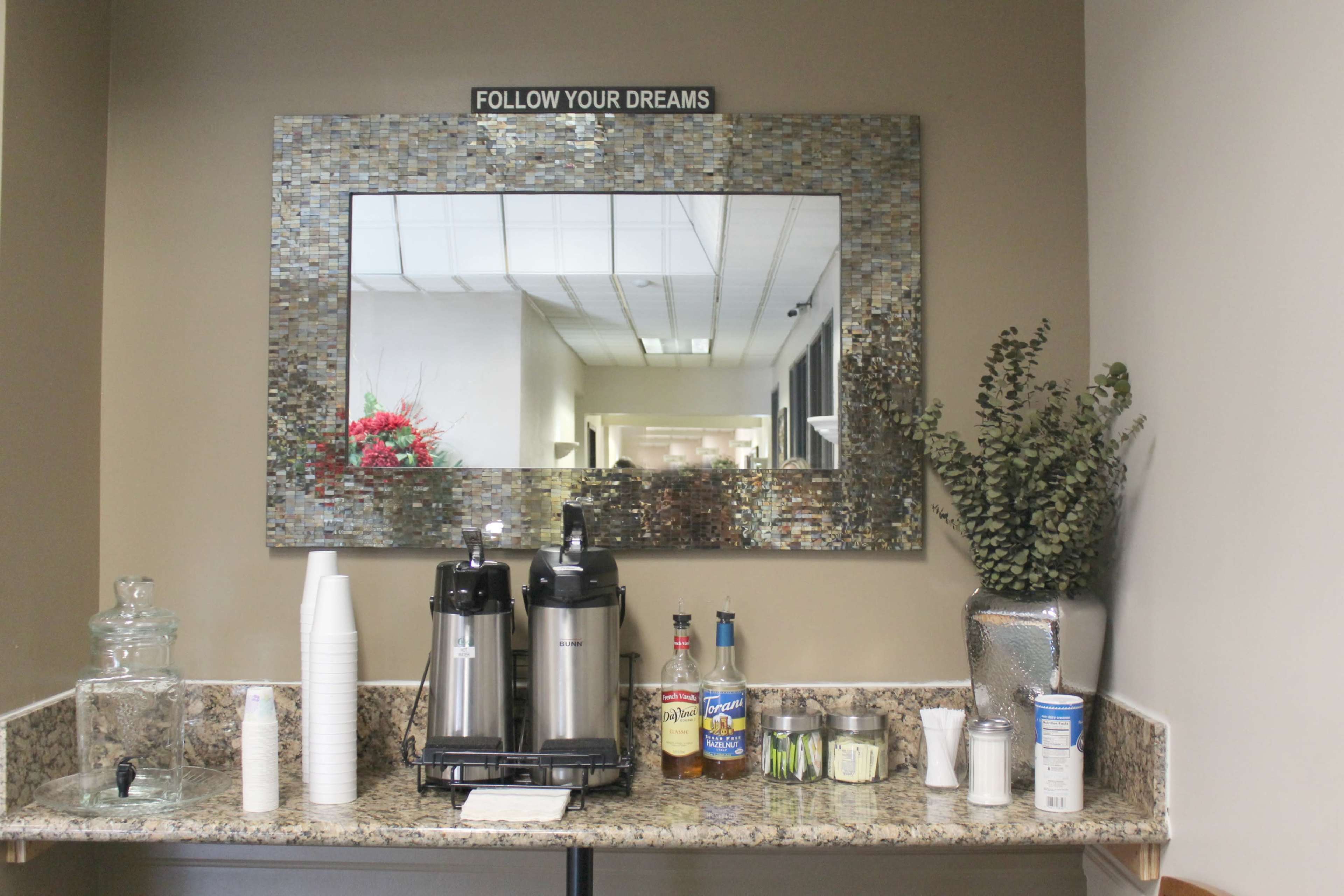 A coffee station featuring two beverage dispensers, cups, and a variety of condiments, situated below a large mirror in a well-lit corridor.