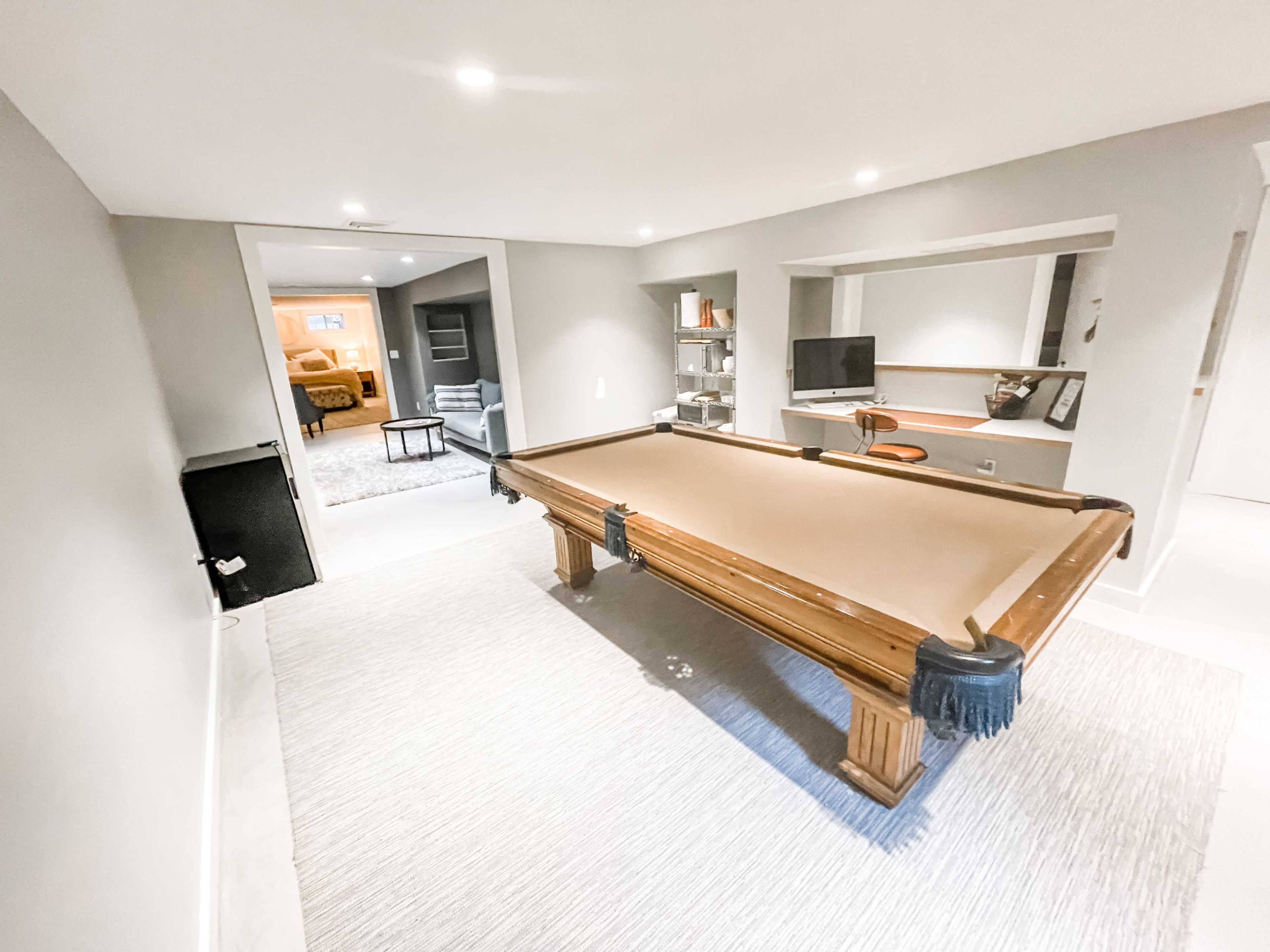 A billiard table occupies the center of a finished basement, with a doorway leading to a living area in the background.