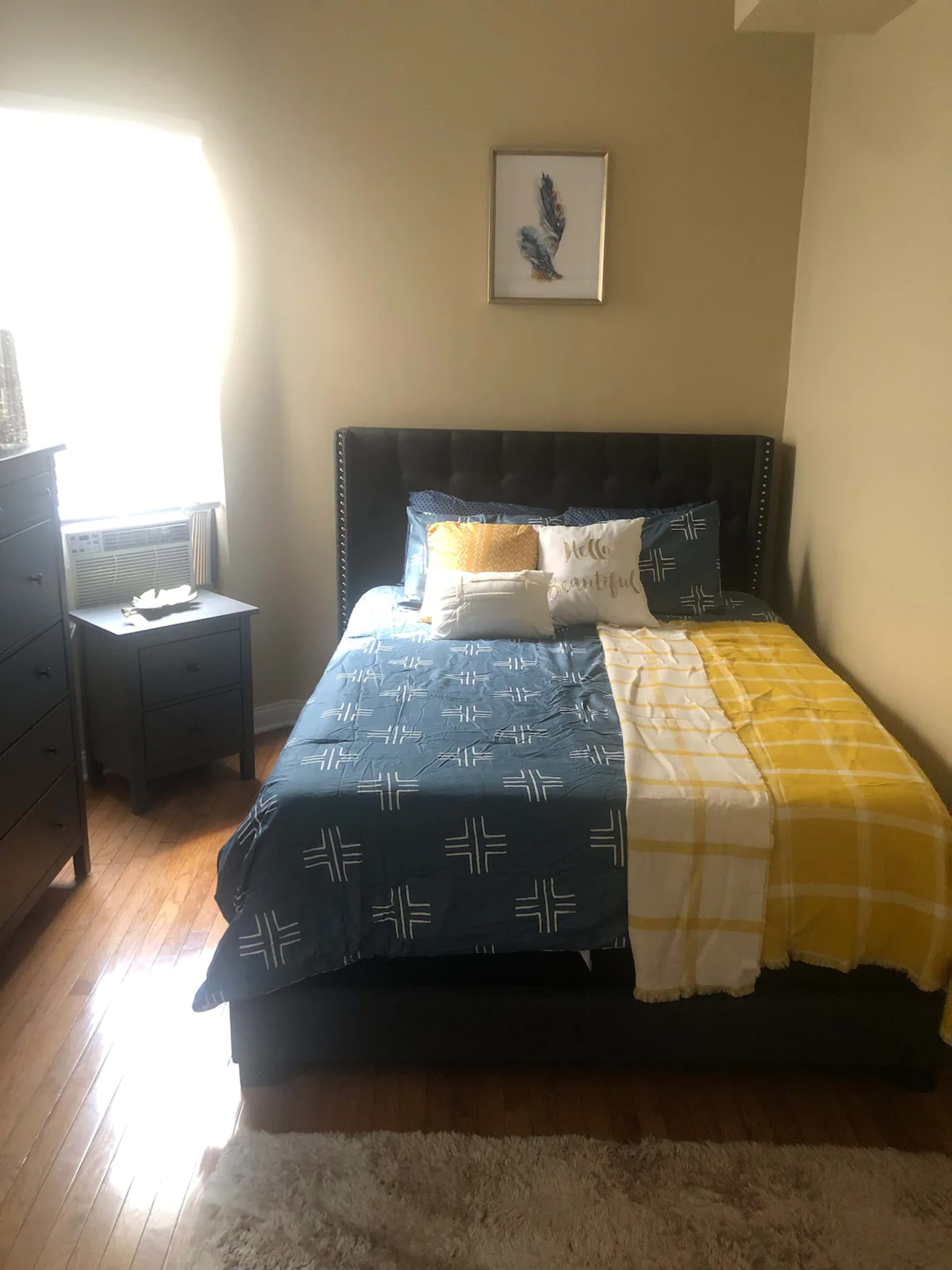 A bedroom featuring a bed with a blue and yellow blanket, a small bedside table, and a dresser under a window with natural light.