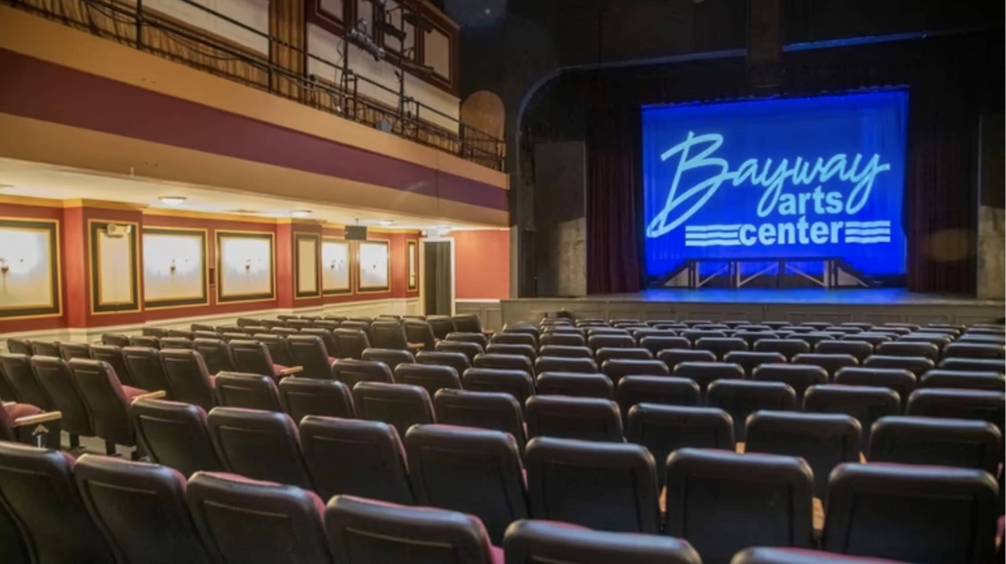 The image depicts a darkened theater with rows of empty seats facing a brightly lit stage featuring a sign that reads "Bayway Arts Center."