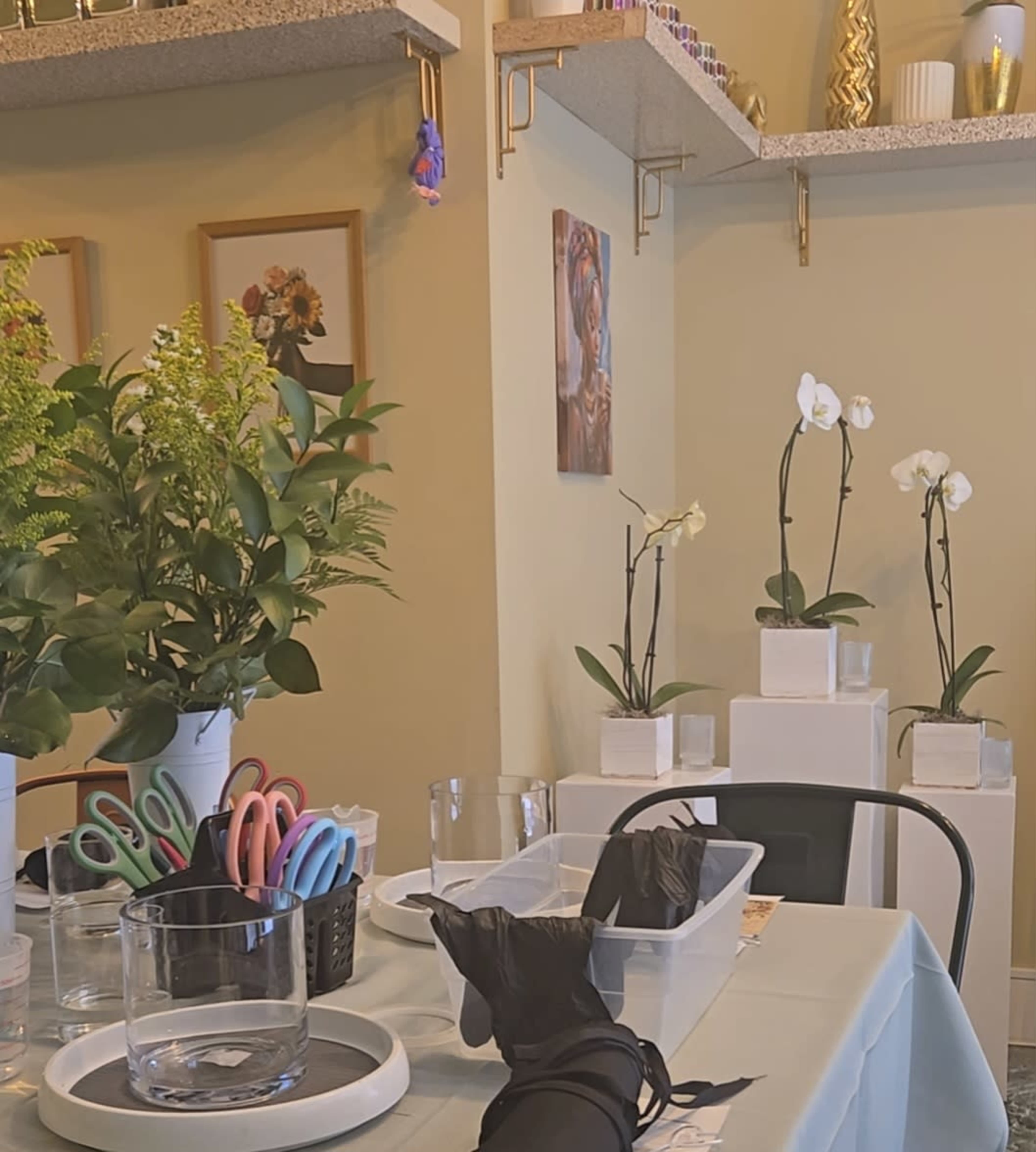 A table is set with various glassware, scissors, and a container under shelves adorned with flowers and decorative items.