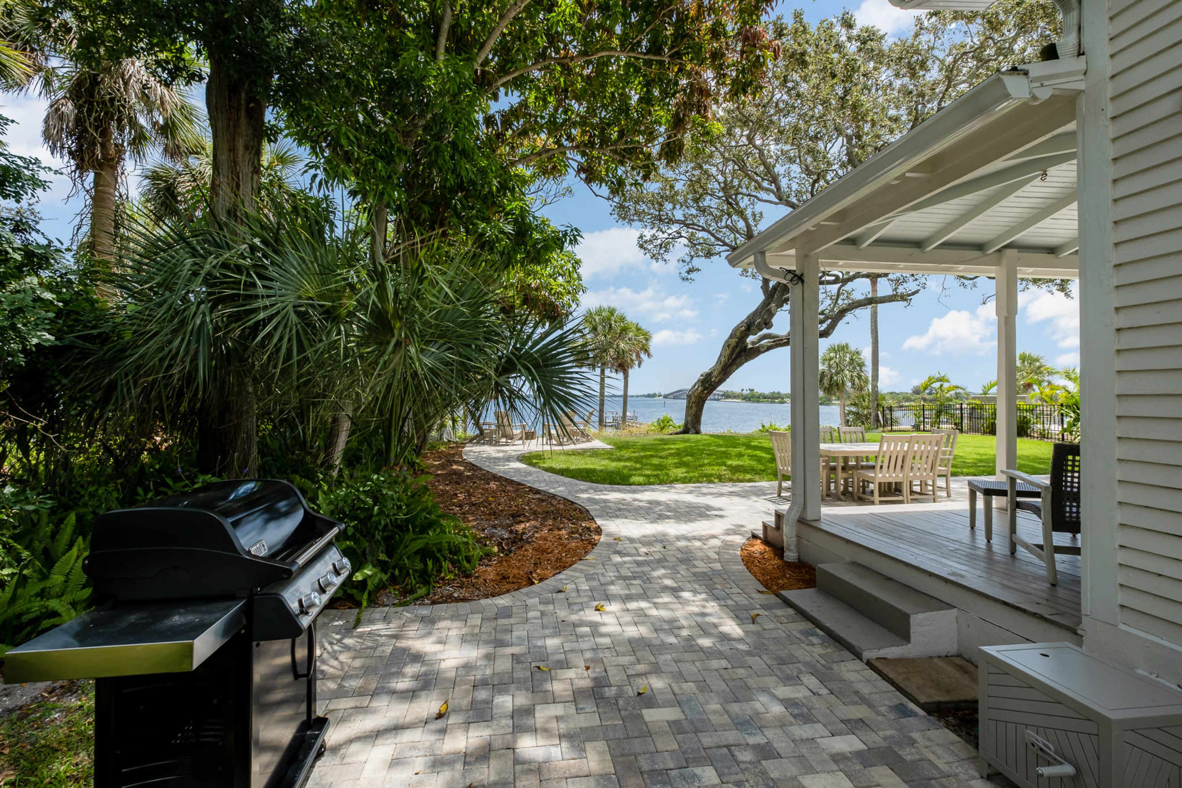 A backyard with a paved pathway leading to a lawn area near the water, featuring a grill and a seating arrangement under a covered porch.