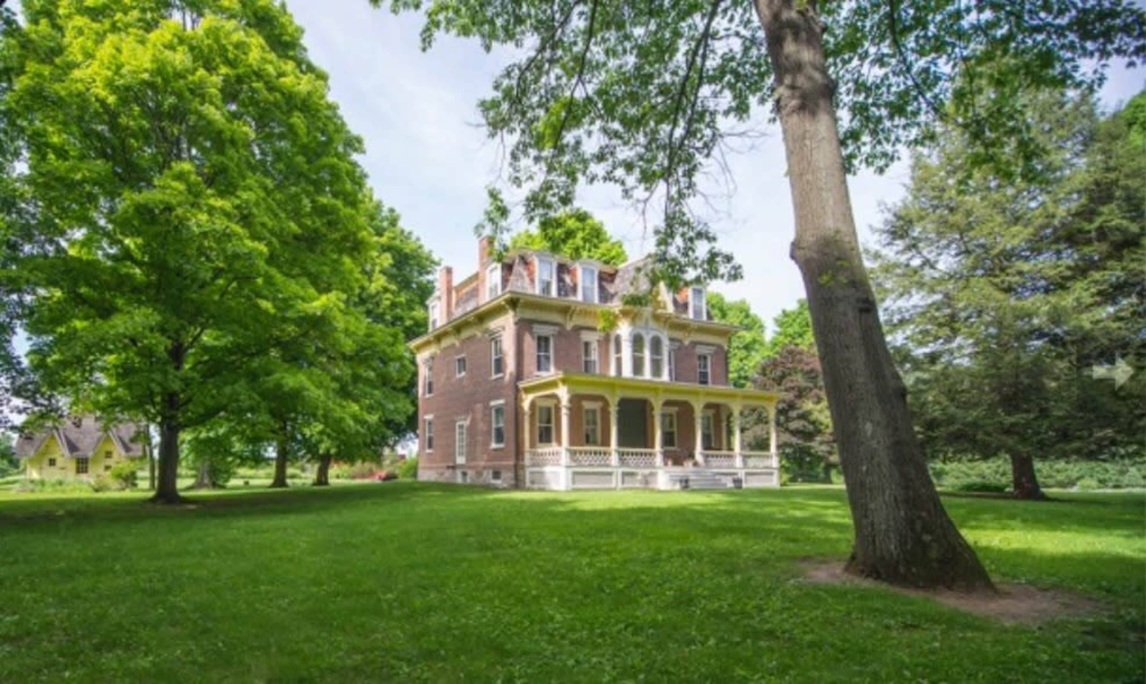 A large brick house with a spacious porch stands surrounded by green grass and trees.
