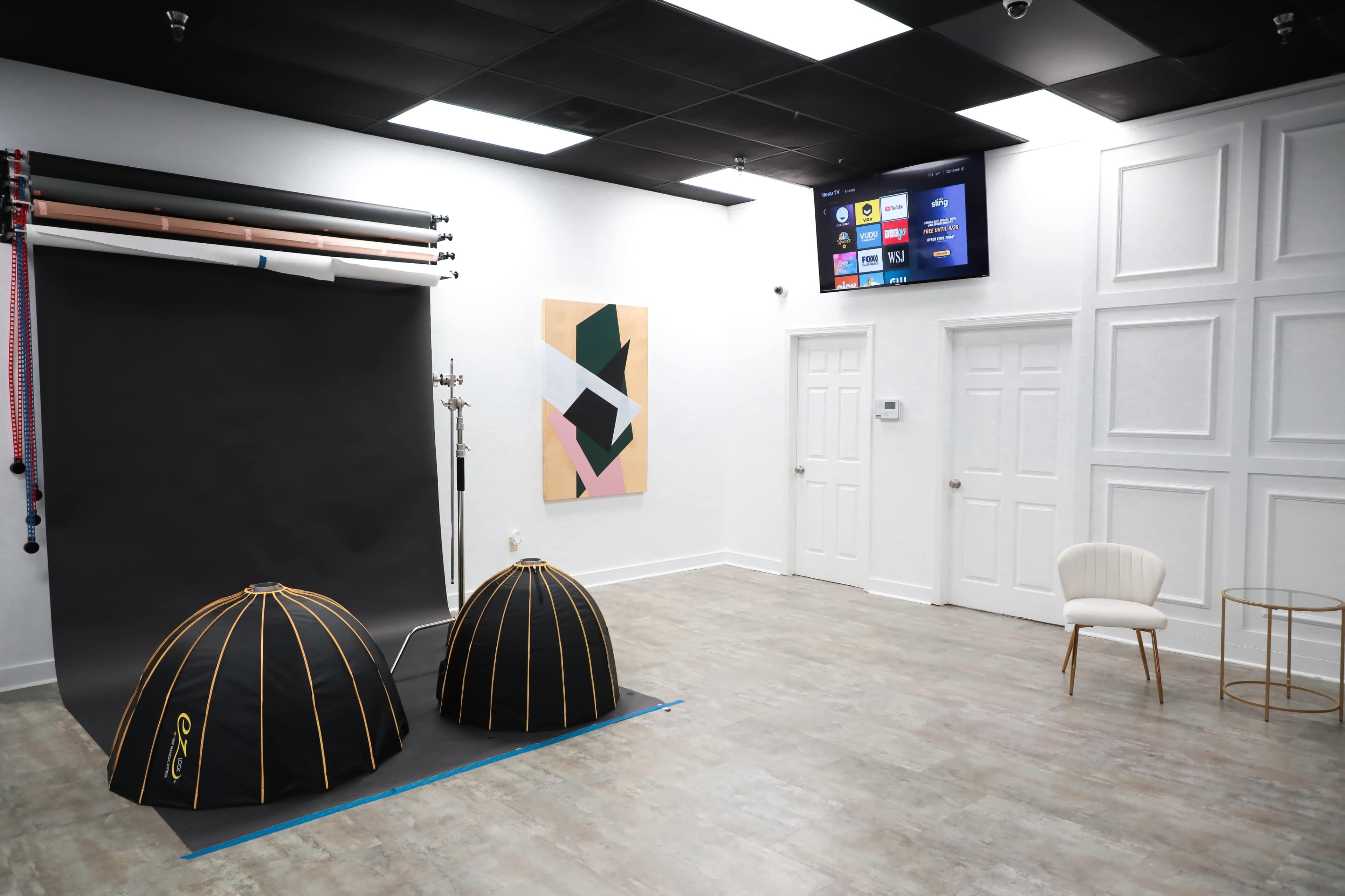 The image shows a photography studio featuring two black diffusers on the floor, a colorful abstract artwork on the wall, a chair, a side table, and a mounted screen displaying information.