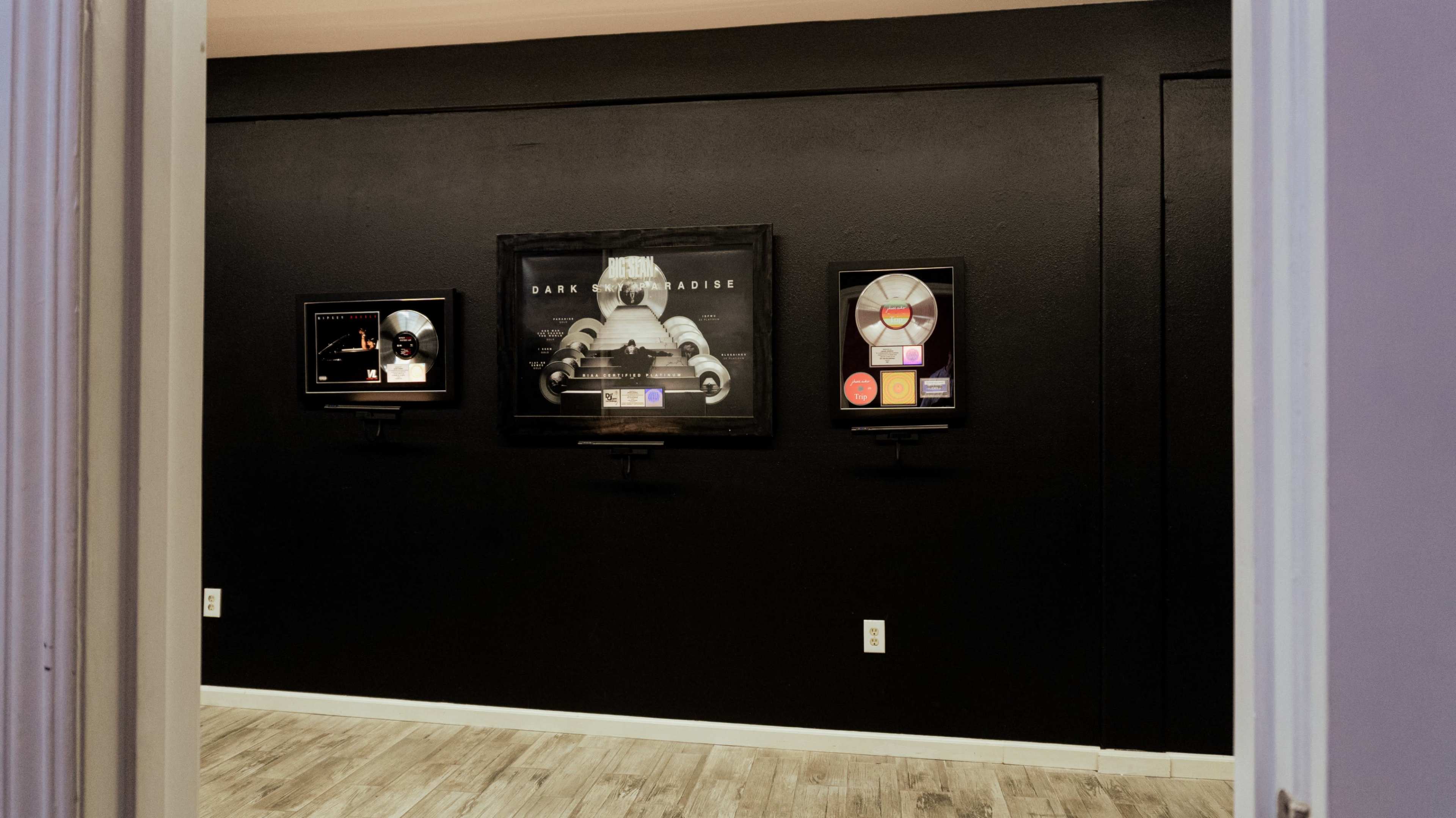 The image shows a dimly lit room with dark walls featuring three framed music awards displayed on the wall.