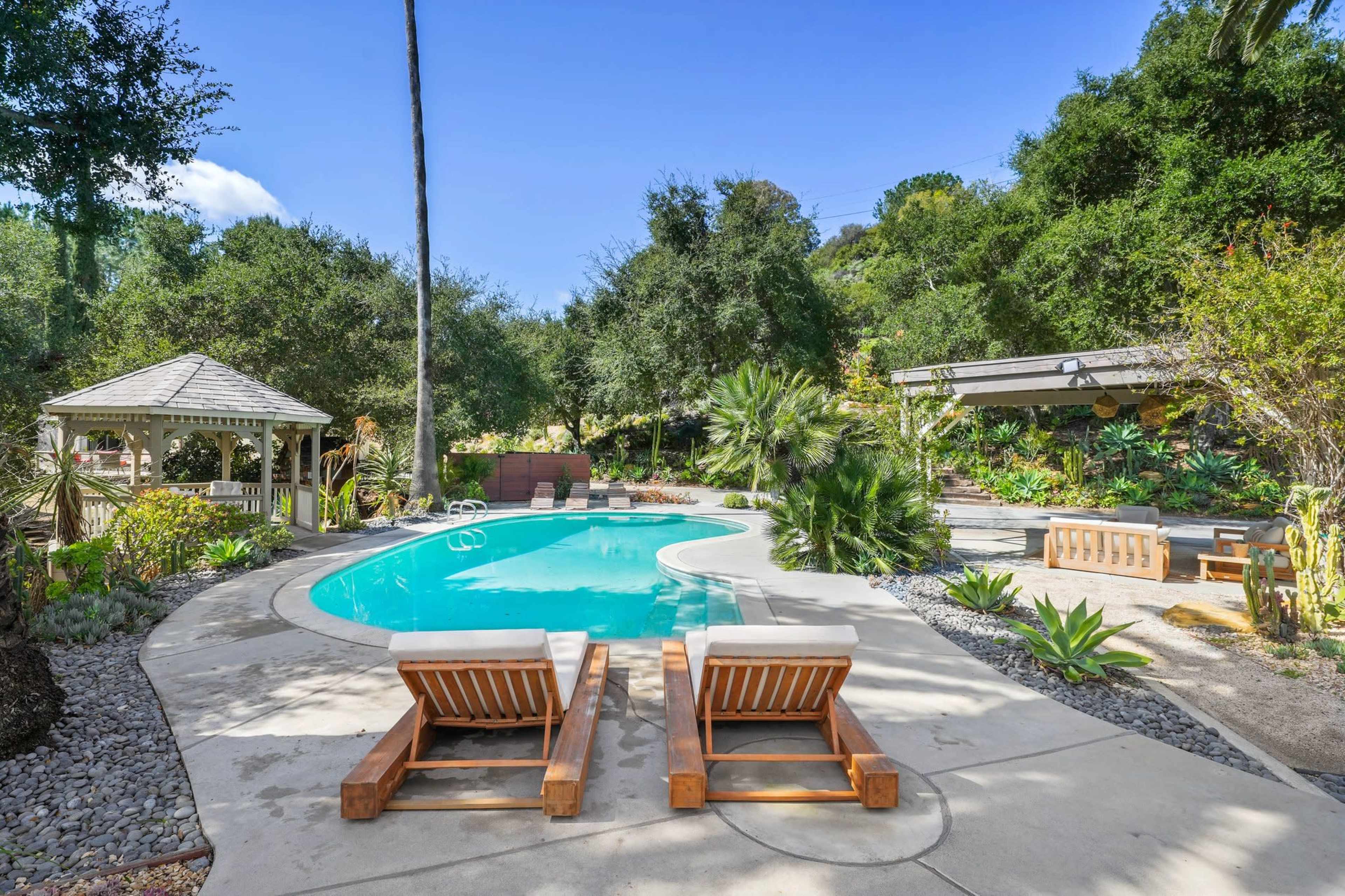 The image shows a landscaped backyard with a swimming pool surrounded by lounge chairs, tropical plants, and a gazebo.