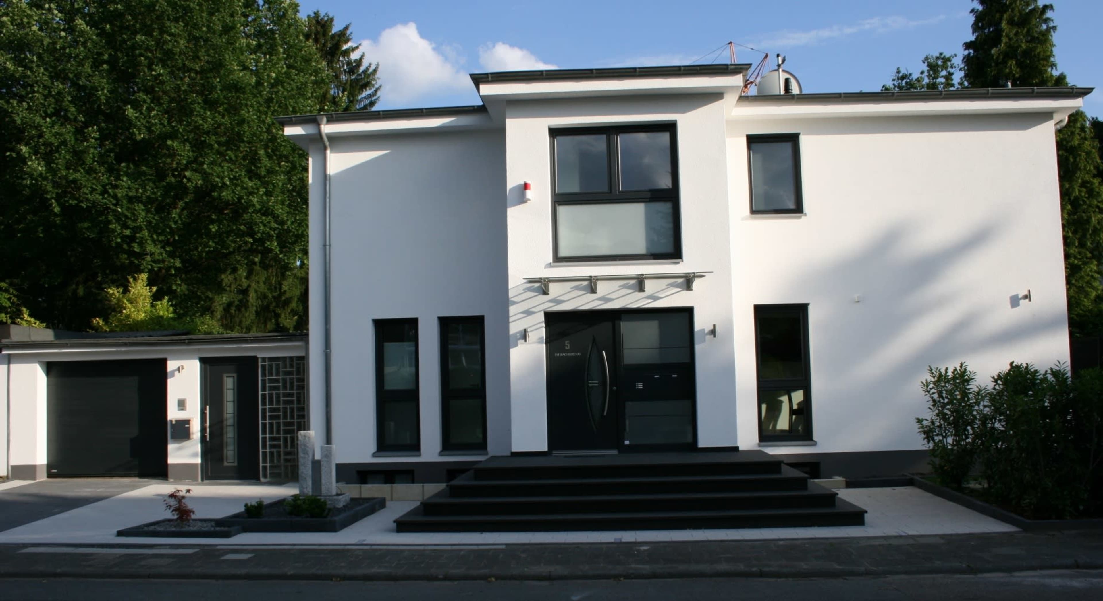 The image shows a modern two-story white house with large black-framed windows, a sleek entrance, and a paved pathway leading to the front door.