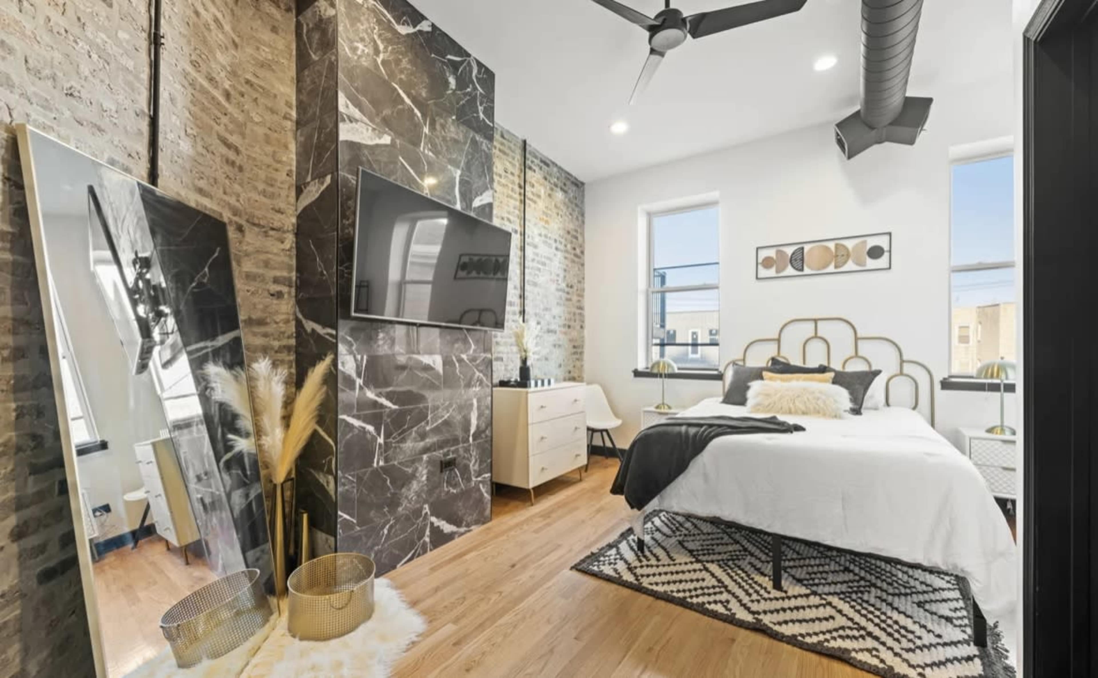 The bedroom features a bed with a black and white color scheme, a large mirror, a television mounted on a marble wall, and wooden flooring.