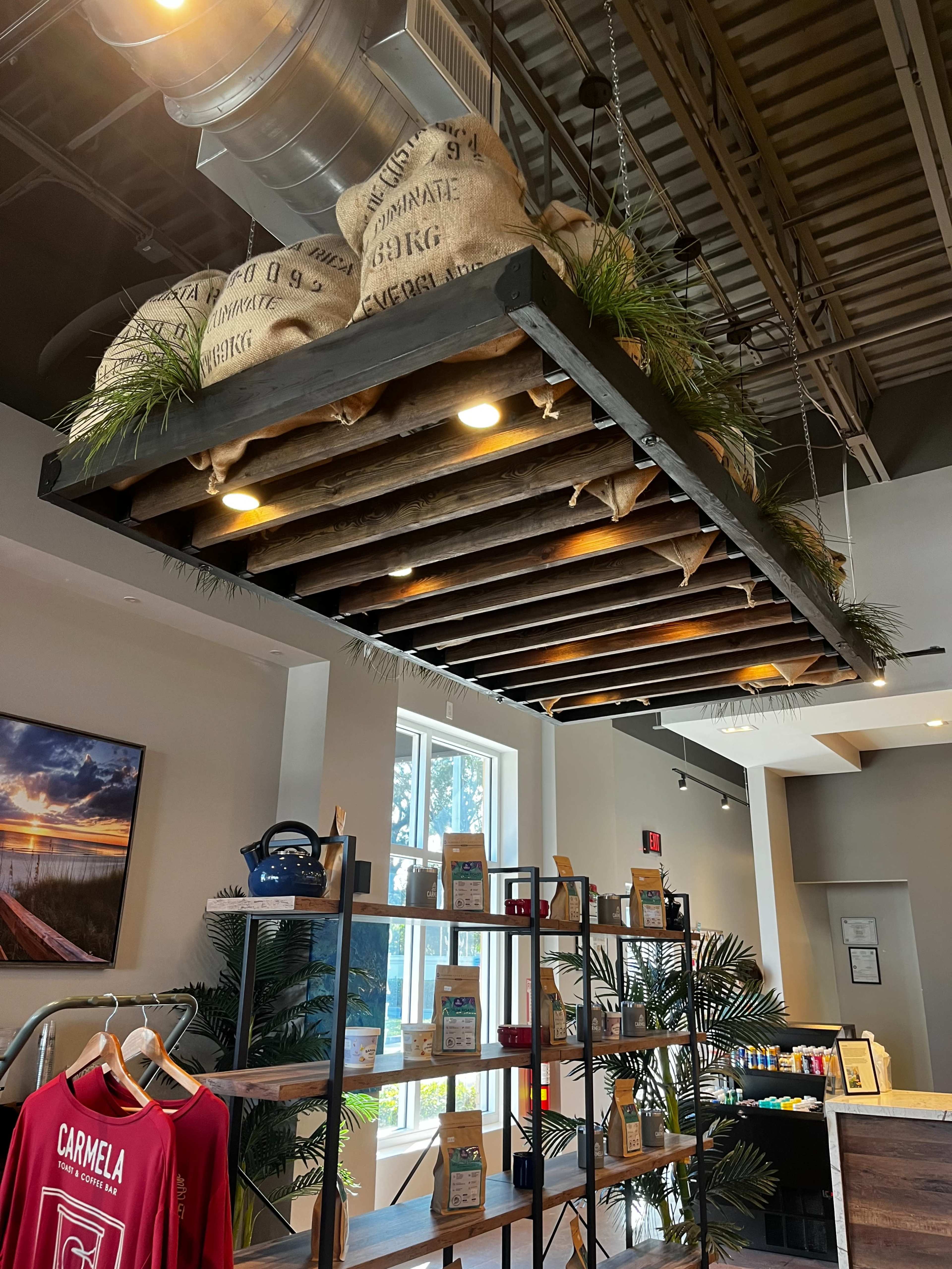The image shows a modern interior space with a wooden ceiling structure adorned with burlap sacks and plants, alongside a shelving unit displaying various products.