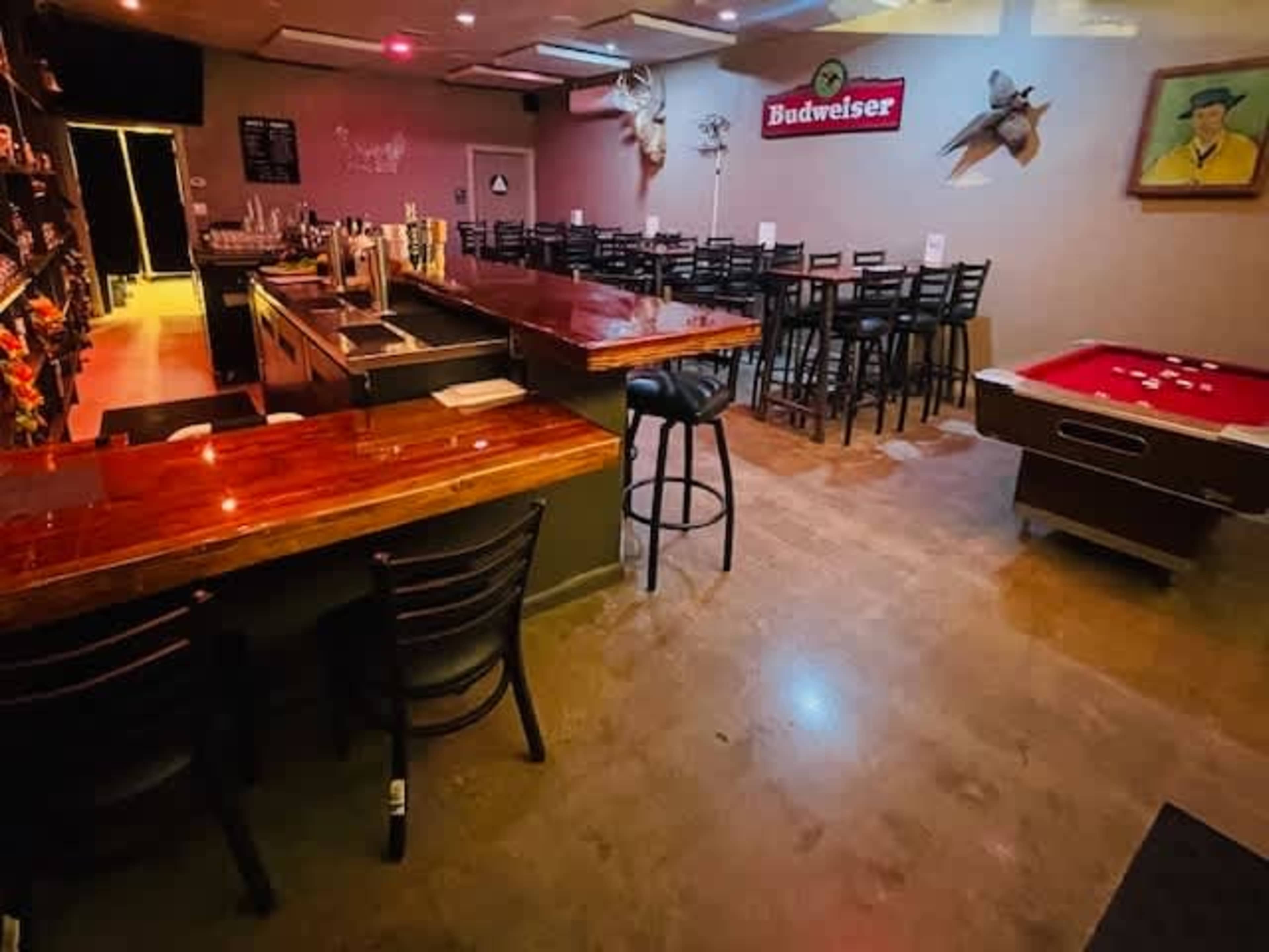 The image shows a bar area with a polished wooden counter and a pool table in a well-lit room featuring a seating area and decorative items on the walls.
