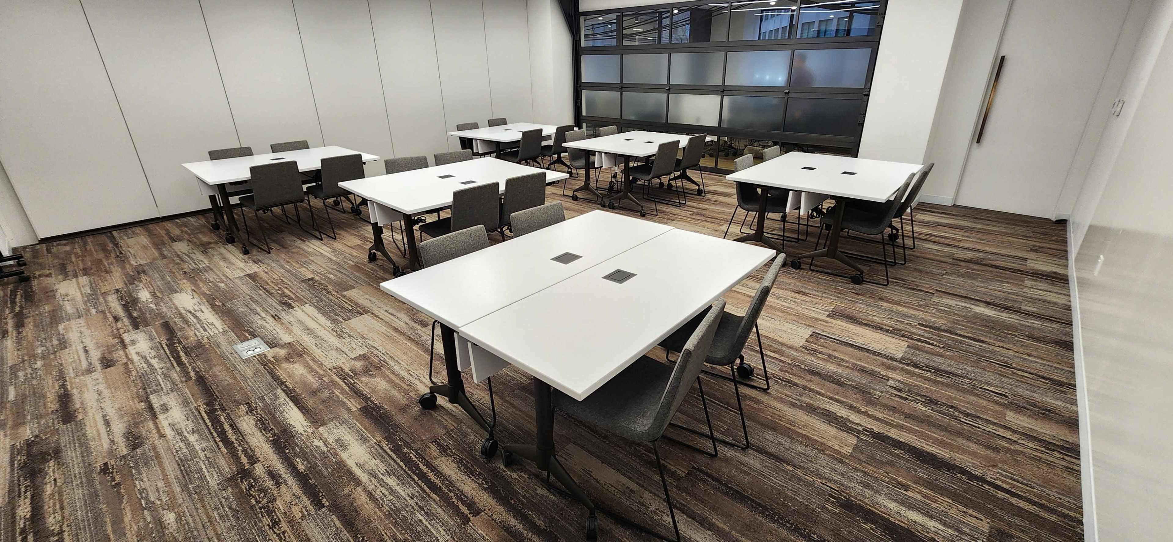 A spacious modern meeting room with multiple white tables and chairs arranged in a grid pattern on a wooden floor.