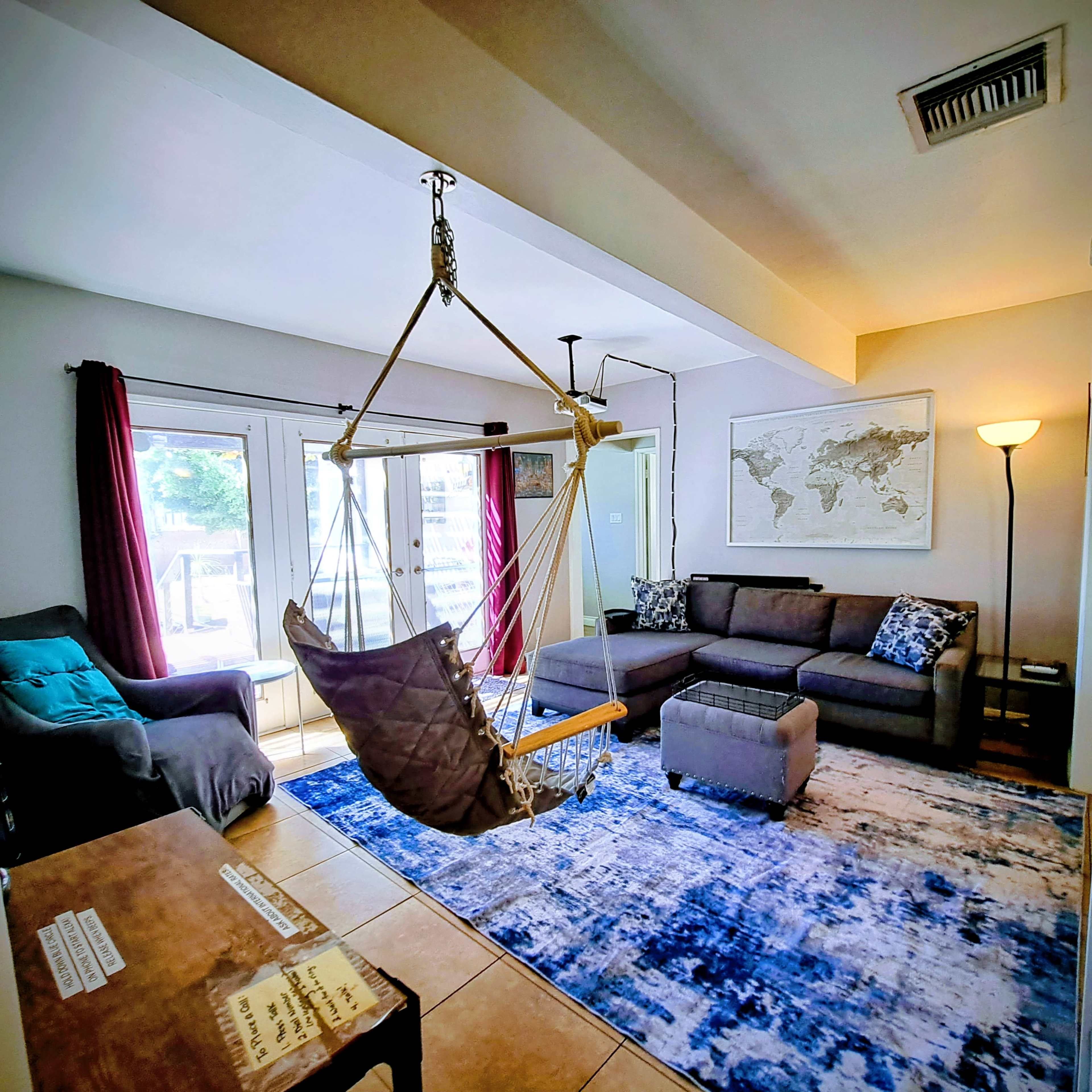 The image shows a living room with a swing chair hanging from the ceiling, a sectional couch, a large area rug, and sliding glass doors leading to an outdoor space.