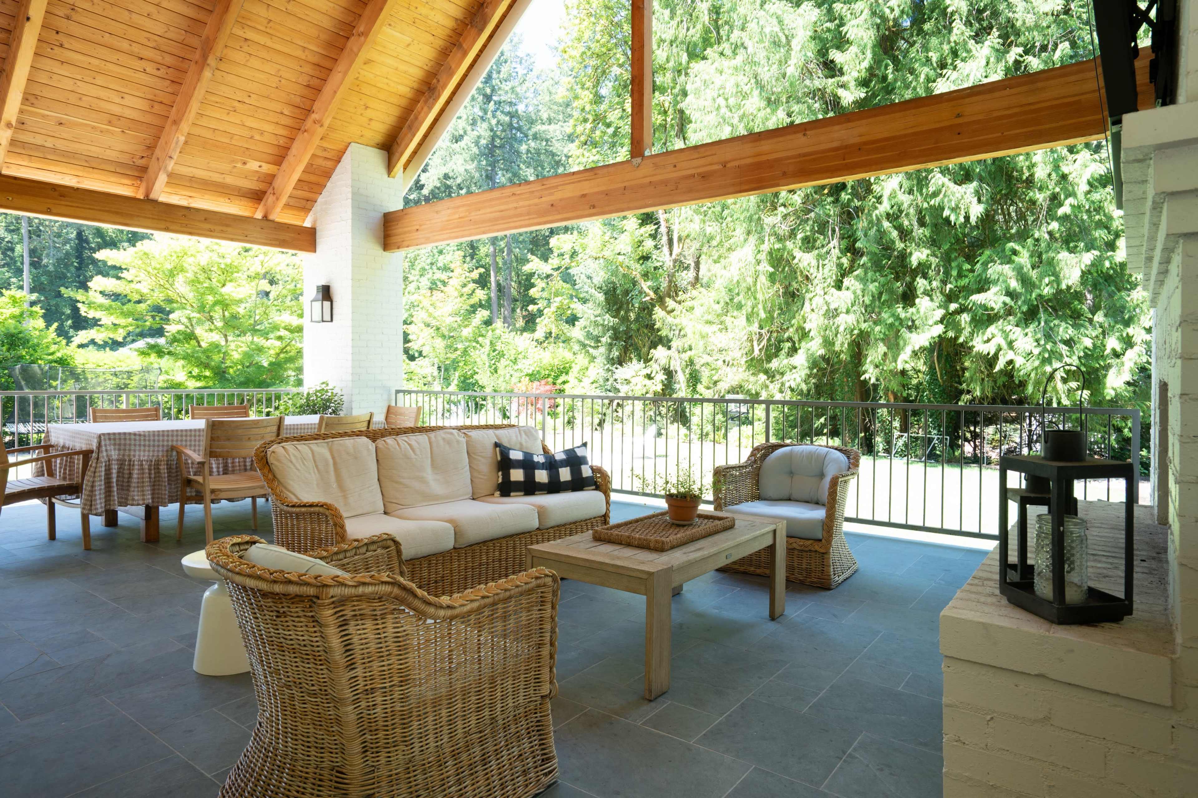 The image shows a spacious outdoor patio featuring a wicker seating arrangement, a wooden table, and surrounded by greenery.