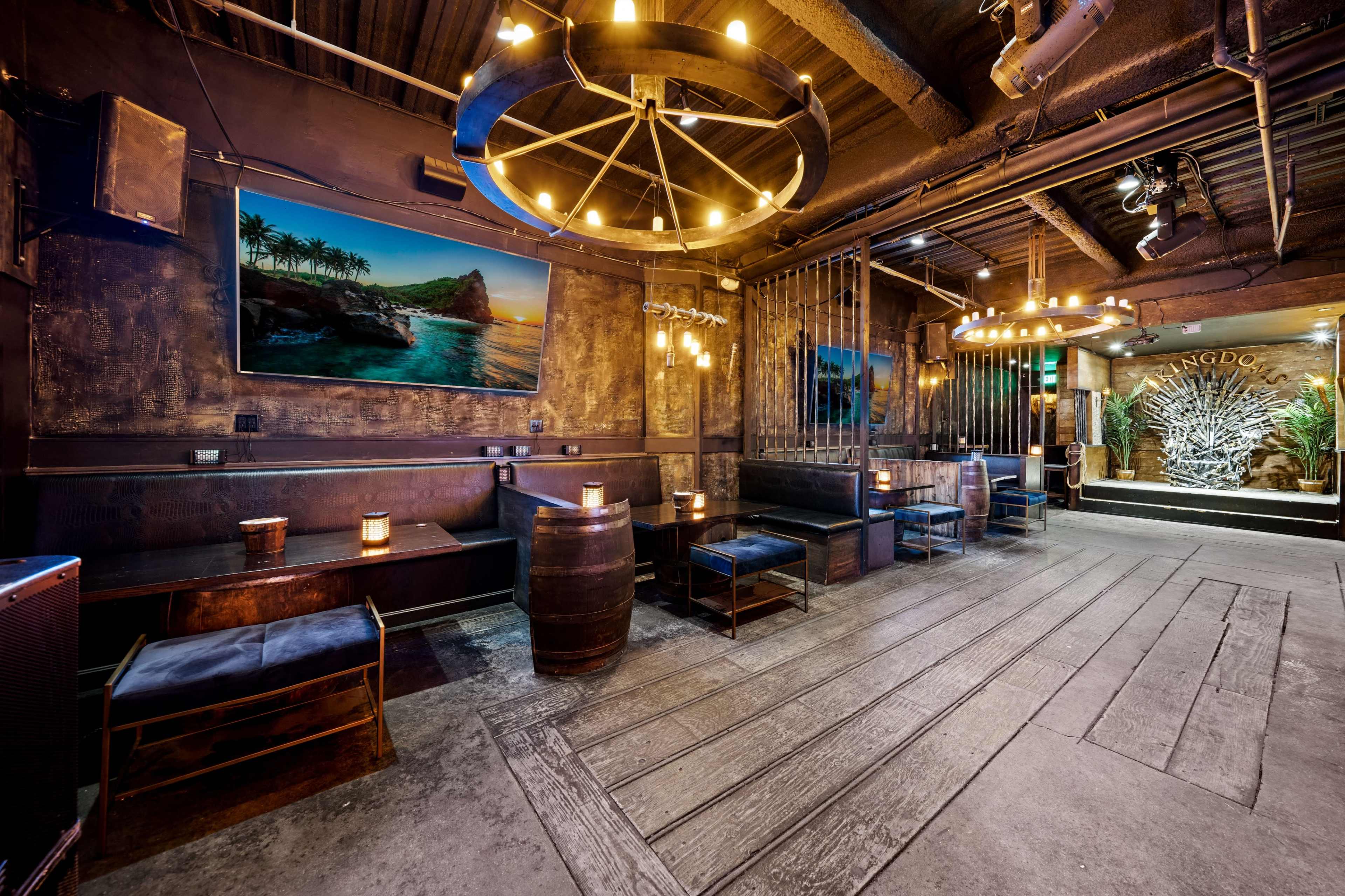 The image shows an interior of a dimly lit bar featuring wooden flooring, seating areas with barrels, and large wall art depicting a coastal scene.