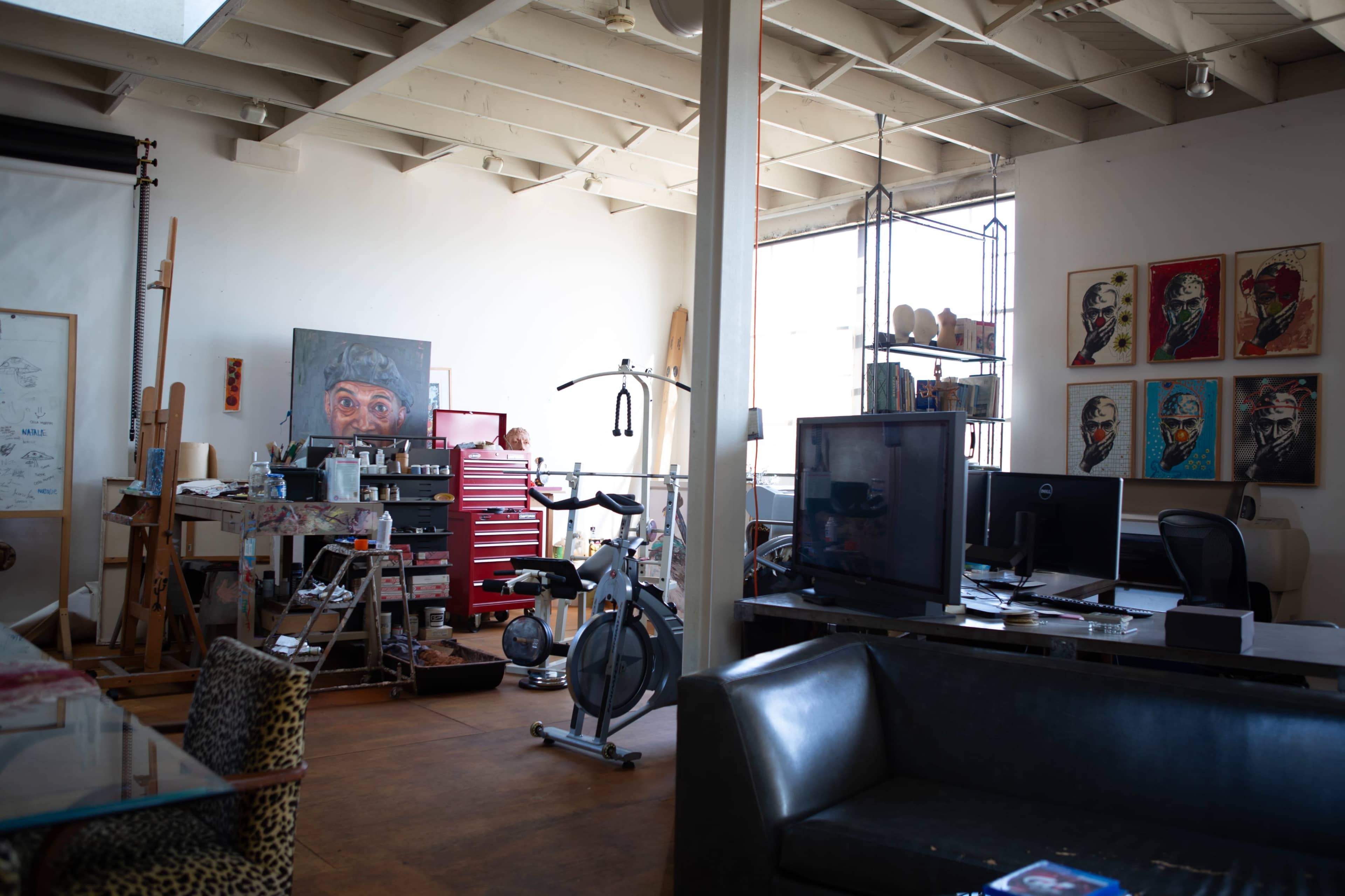 The image shows an artist's studio featuring easels, art supplies, a workout bike, and various artworks on the walls.
