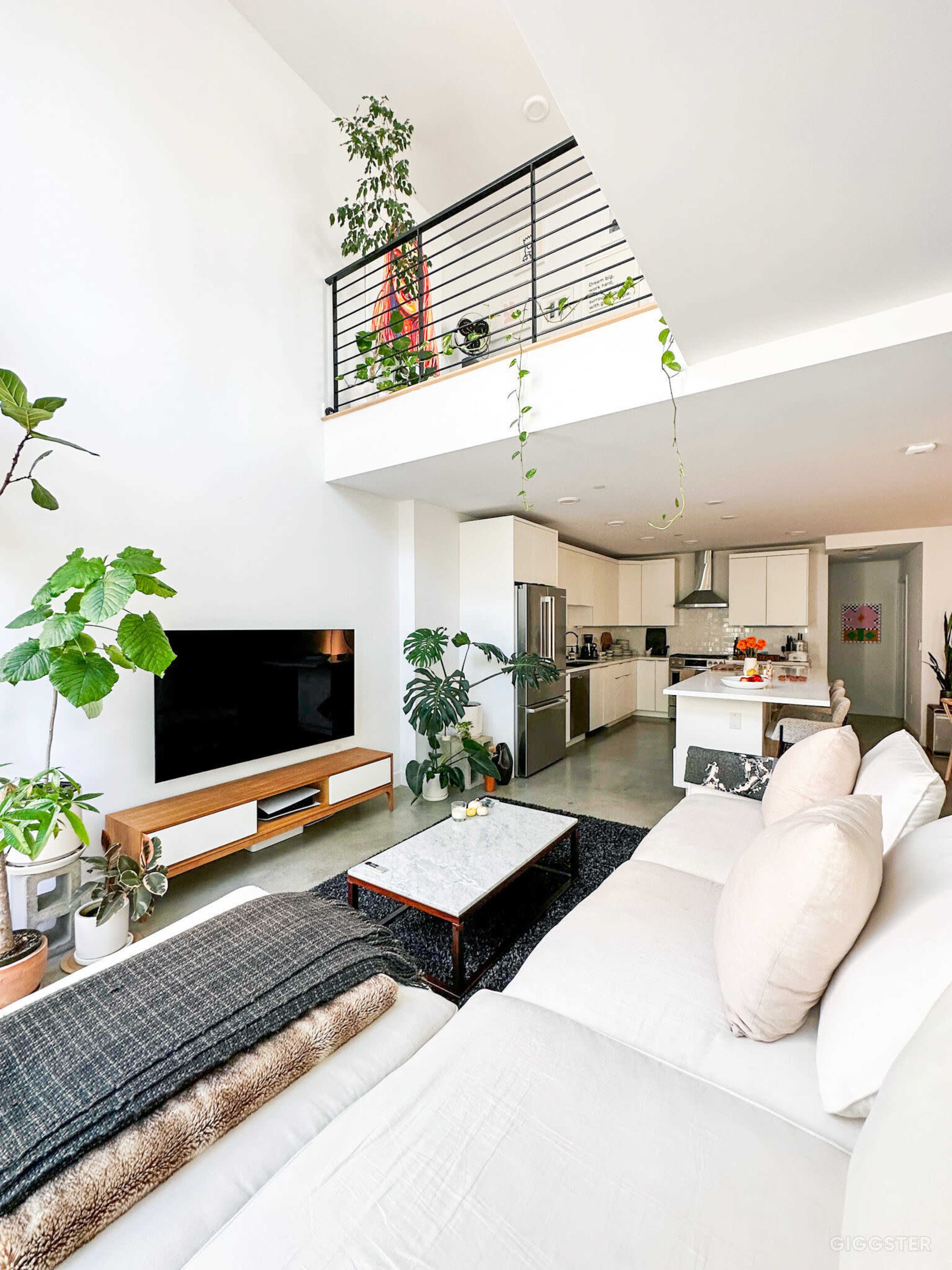 A modern living room with a large sectional sofa, a coffee table, indoor plants, and an open kitchen area visible in the background.