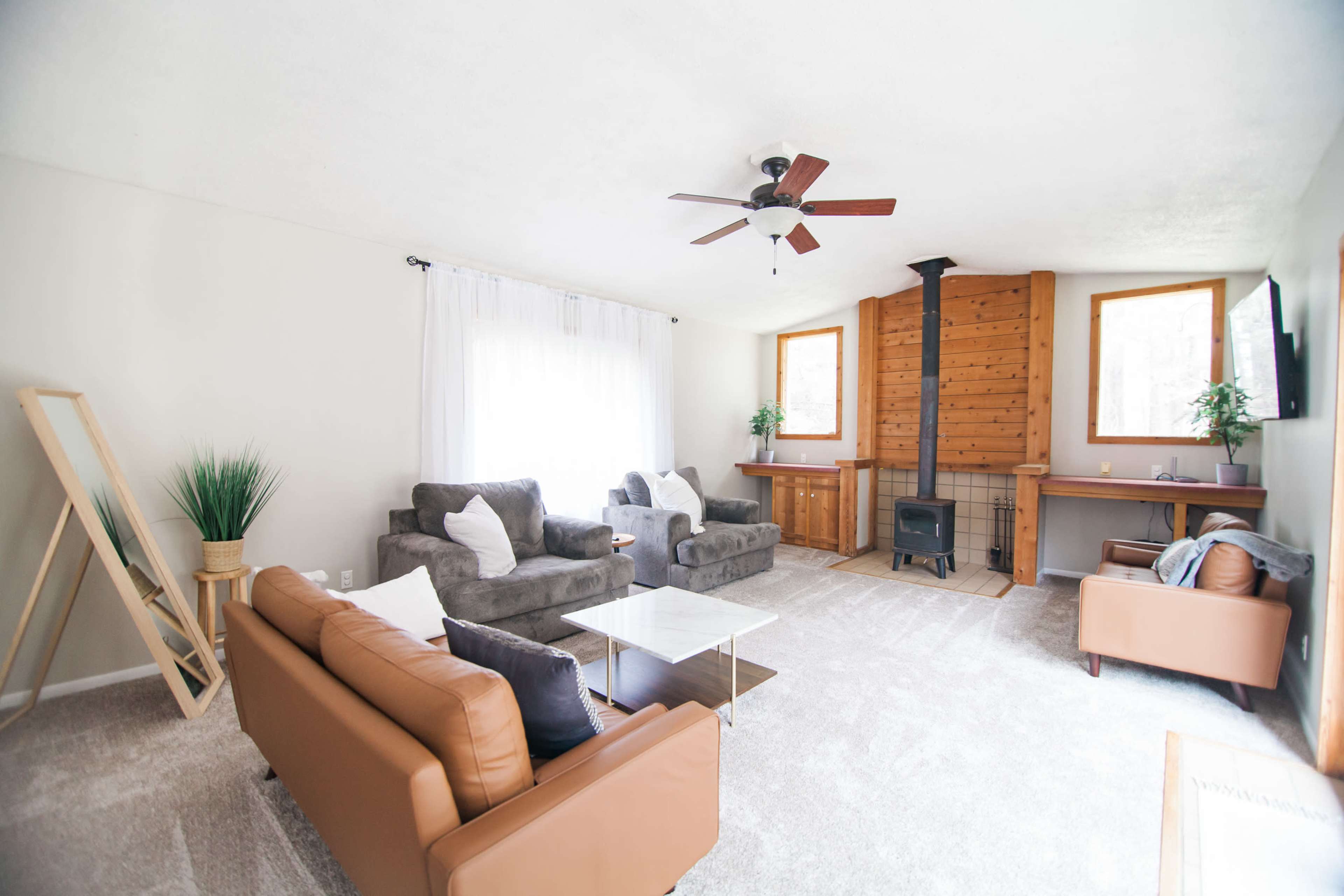 The living room features two seating areas with gray sofas and a brown leather couch, a wood-paneled wall, and a central wood stove, complemented by large windows and a light-colored carpet.