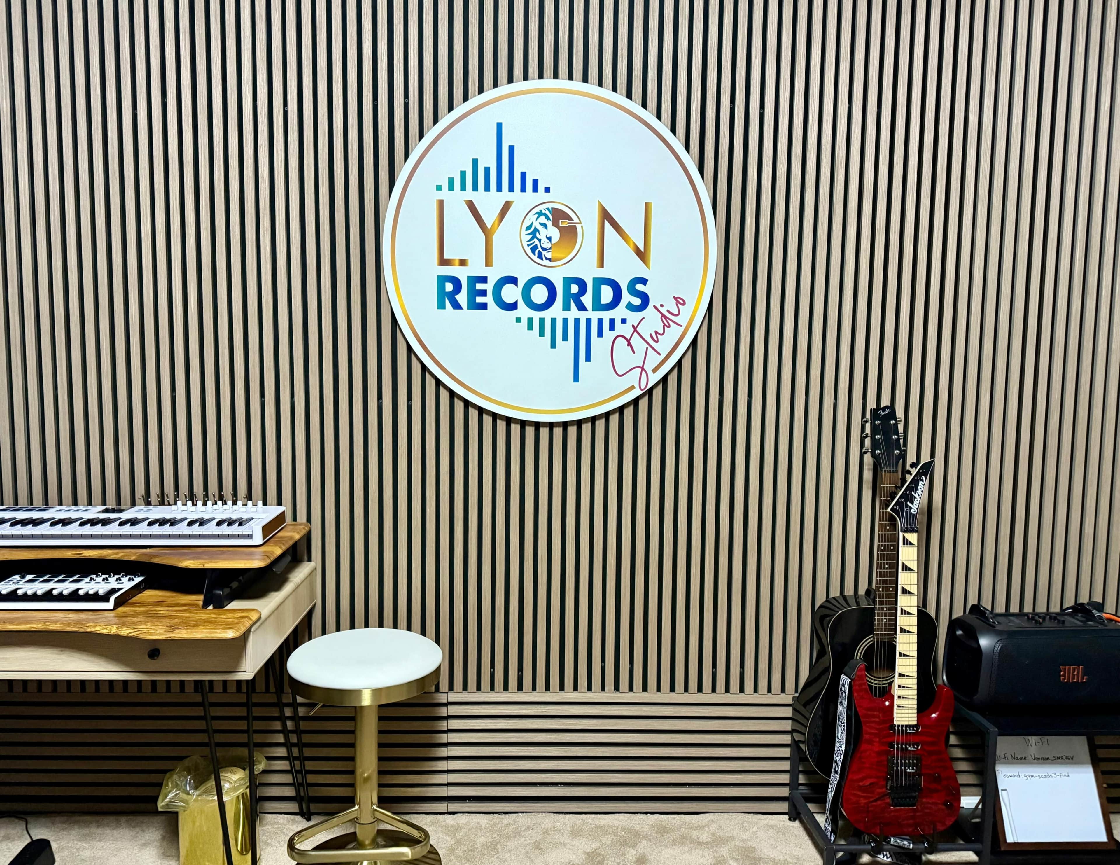 The image shows a music studio wall featuring a circular sign that reads "LYON RECORDS" along with a keyboard, a stool, and an electric guitar positioned next to a speaker.