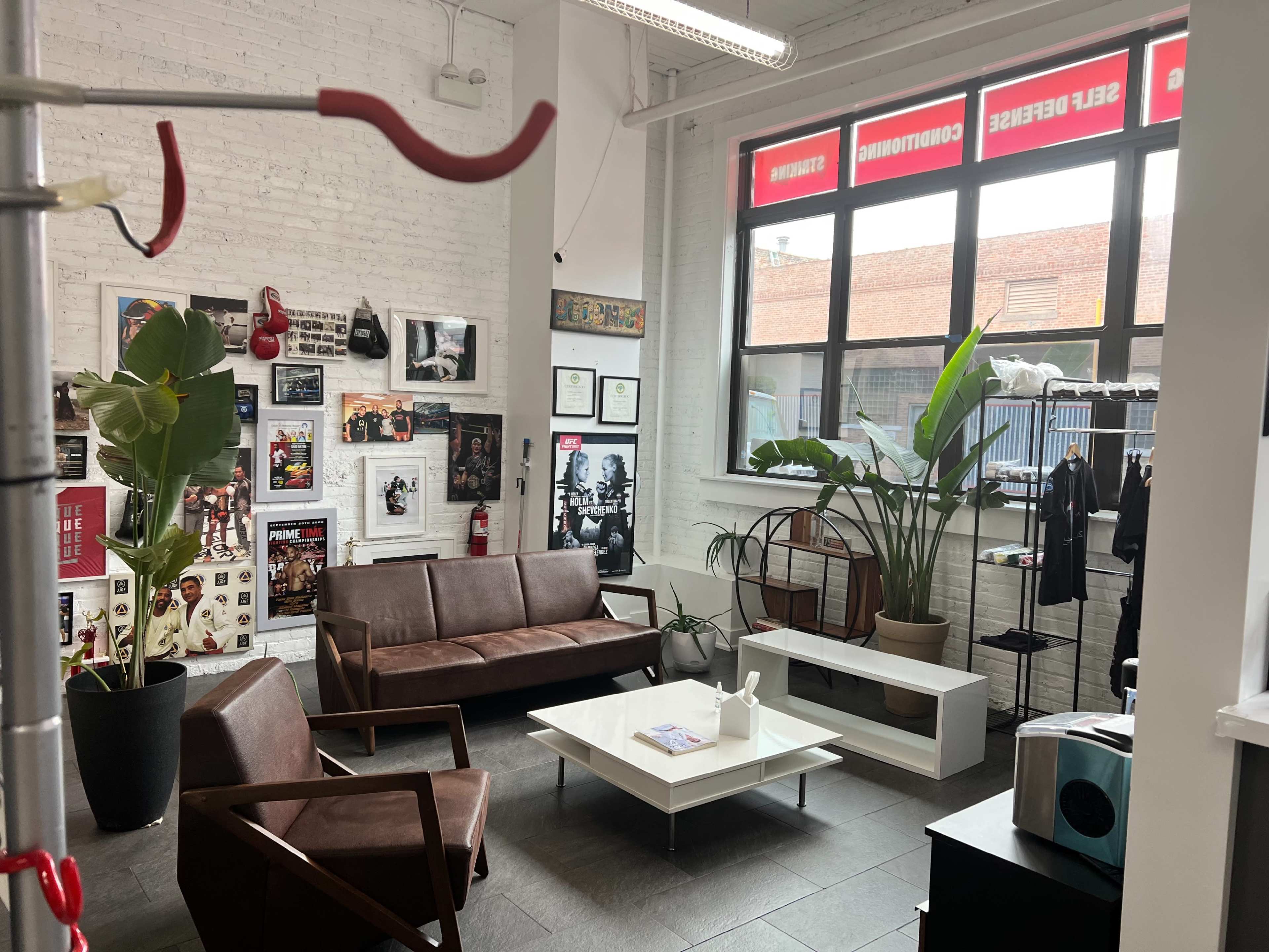 A modern waiting area featuring brown leather couches, a white coffee table, potted plants, and framed photographs on the walls, with large windows overlooking an outdoor space.