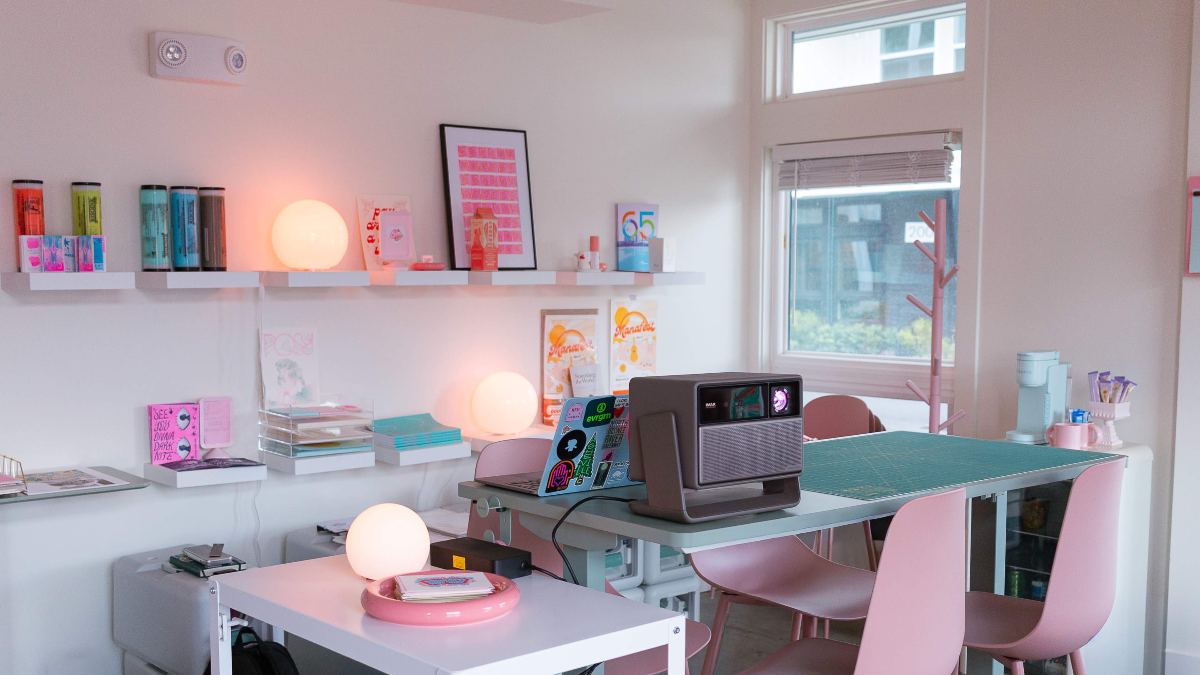 The image depicts a modern workspace featuring a table with a laptop, pastel-colored chairs, and shelves displaying various decorative items and stationary.