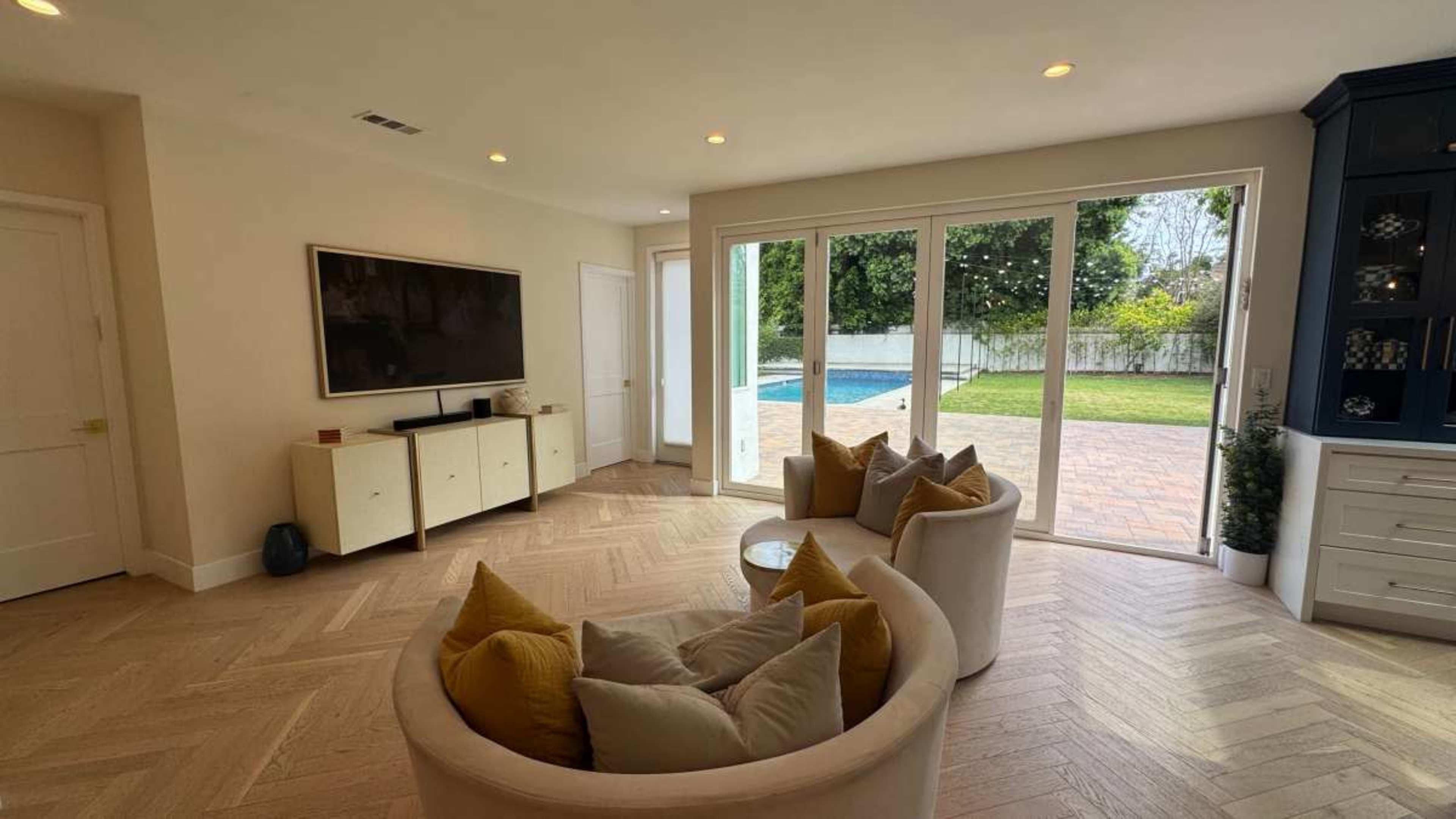 The image shows a living room with a television on the wall, two circular sofas with pillows, and large glass doors leading to a backyard with a pool.