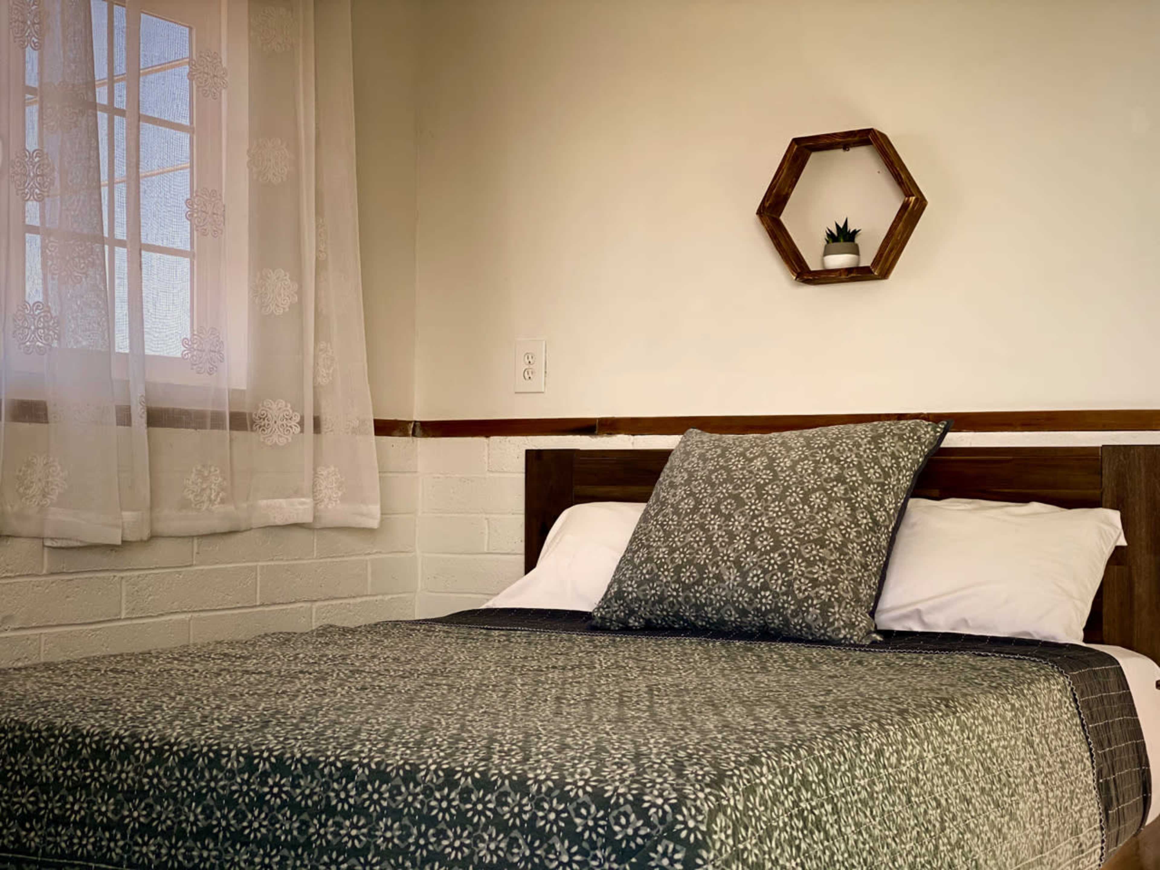 A neatly made bed with a patterned quilt and a large pillow is positioned against a wall with a small hexagonal shelf holding a plant.