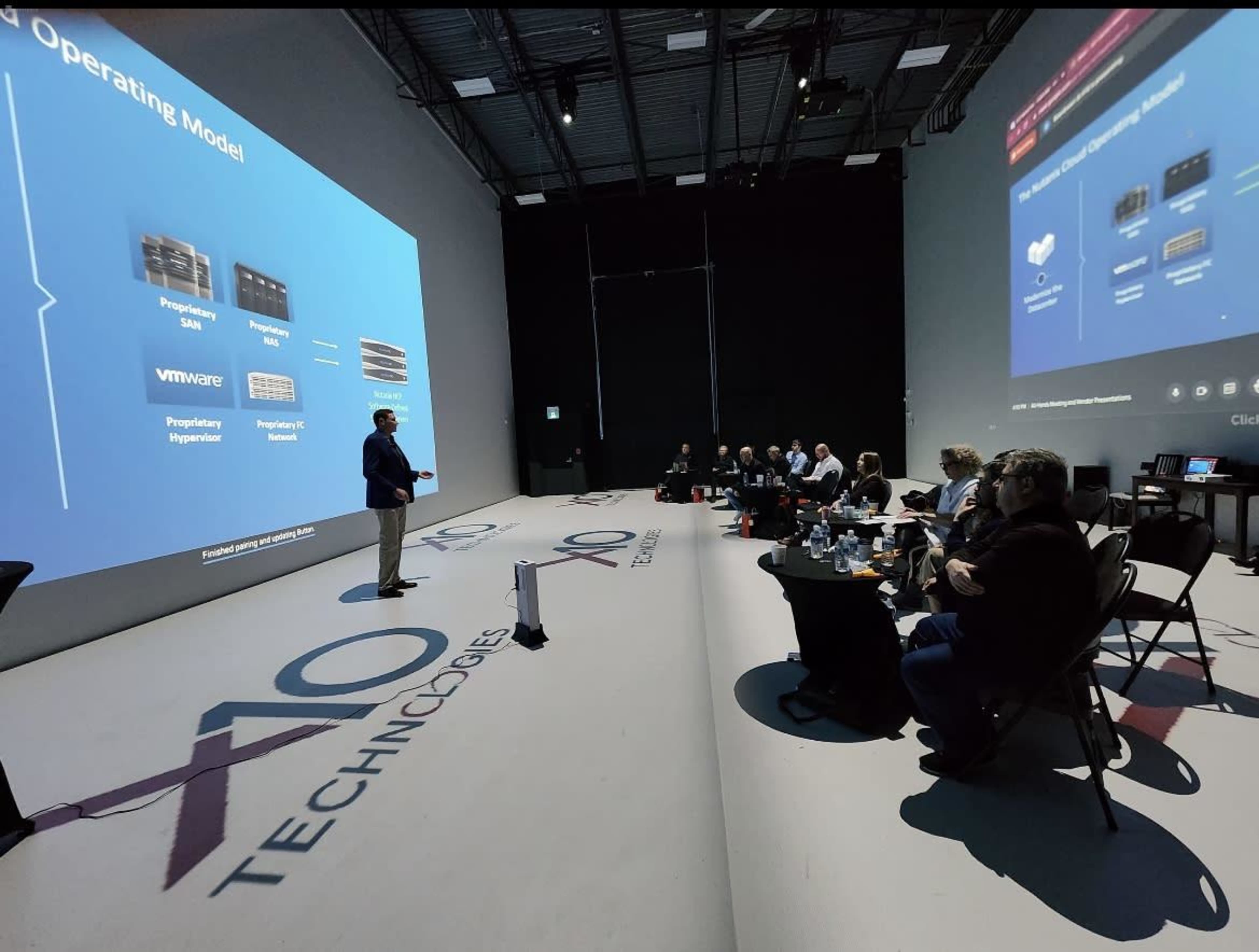 A speaker presents information on a large screen to an audience seated in a dimly lit conference room.