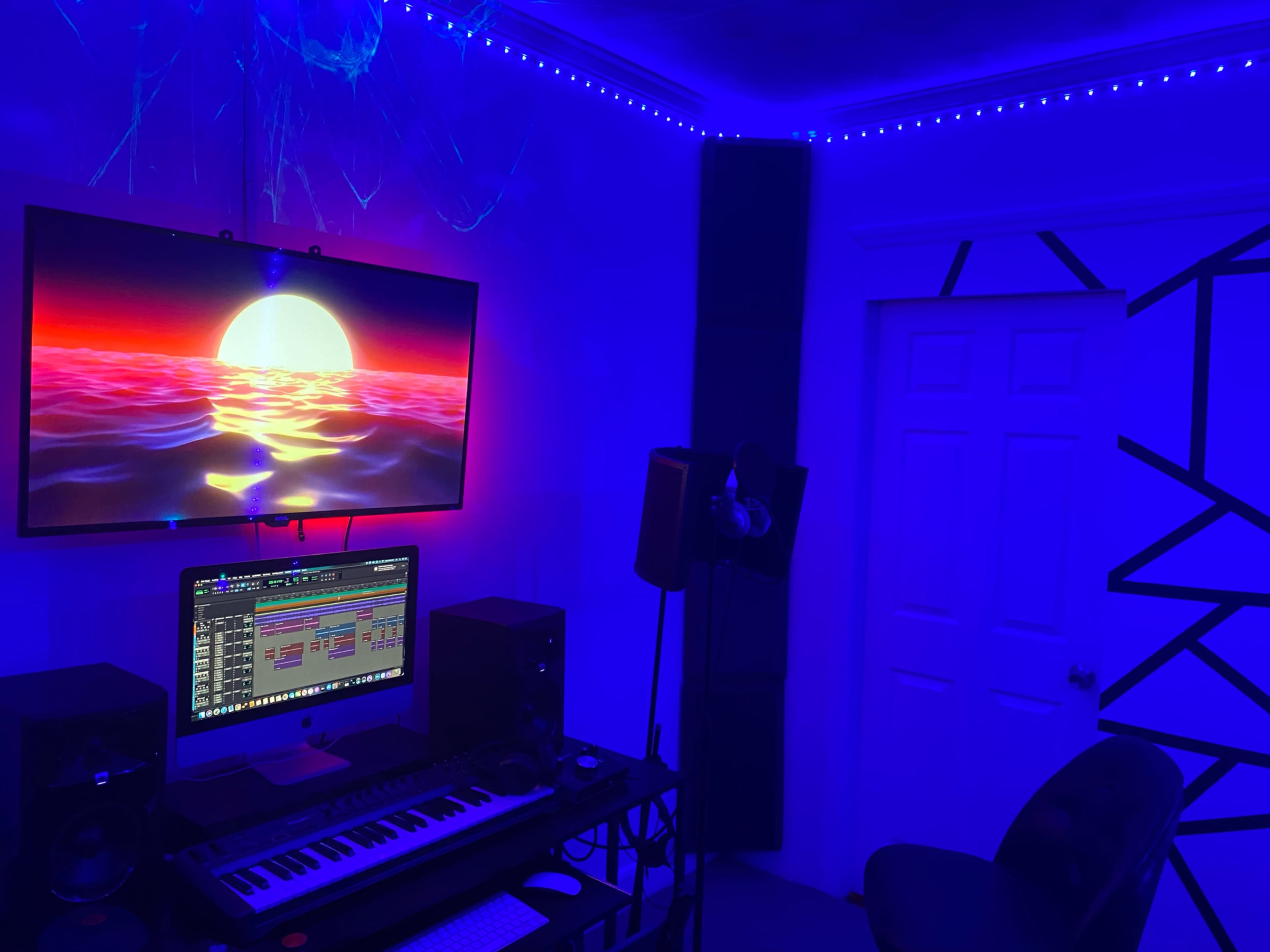A music production setup with a computer, MIDI keyboard, and speakers in a room illuminated by blue LED lights and a large screen displaying a sunset over water.