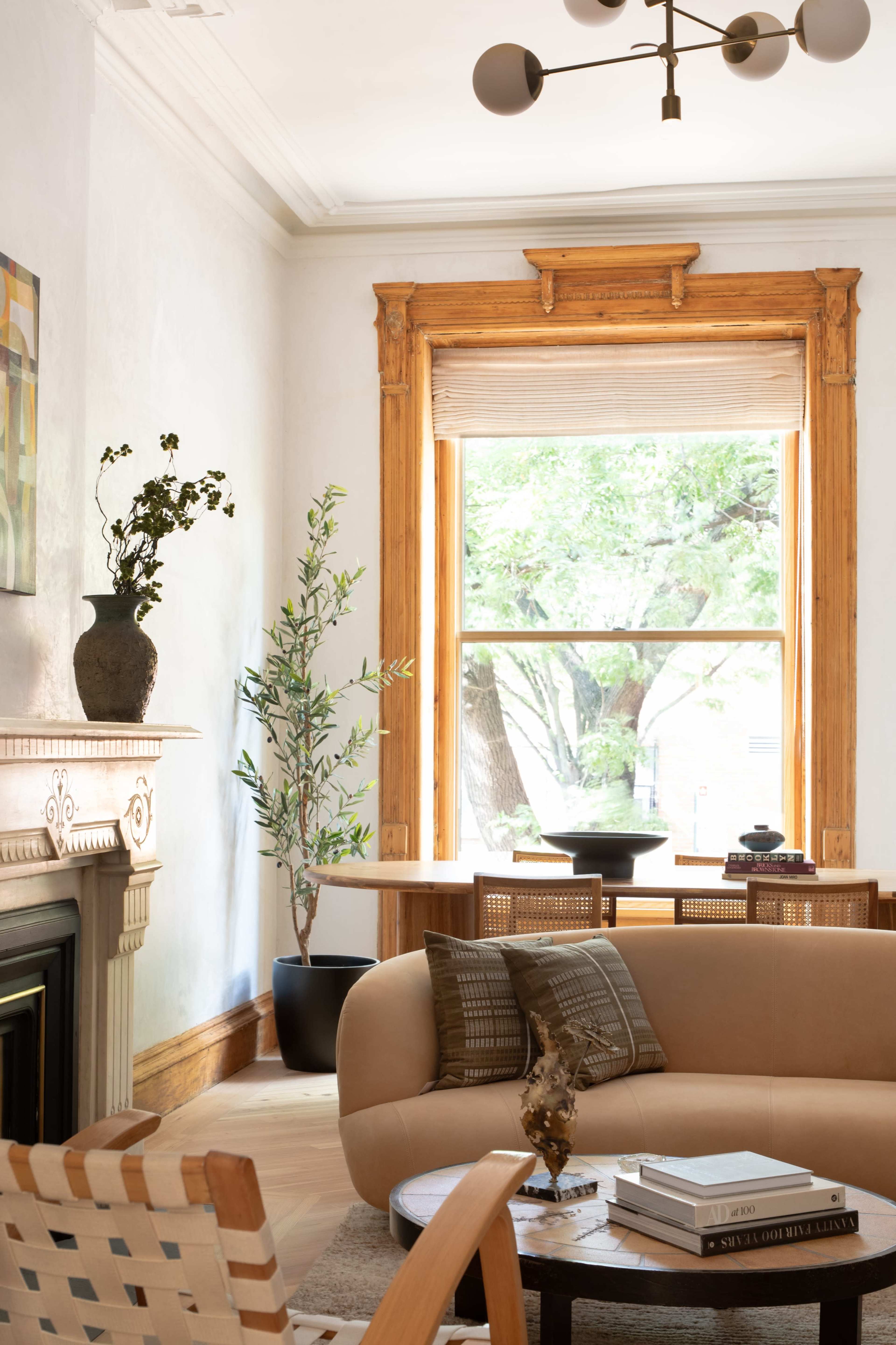 The image shows a cozy living room with a large window, decorative plants, and modern furniture arranged around a central coffee table.