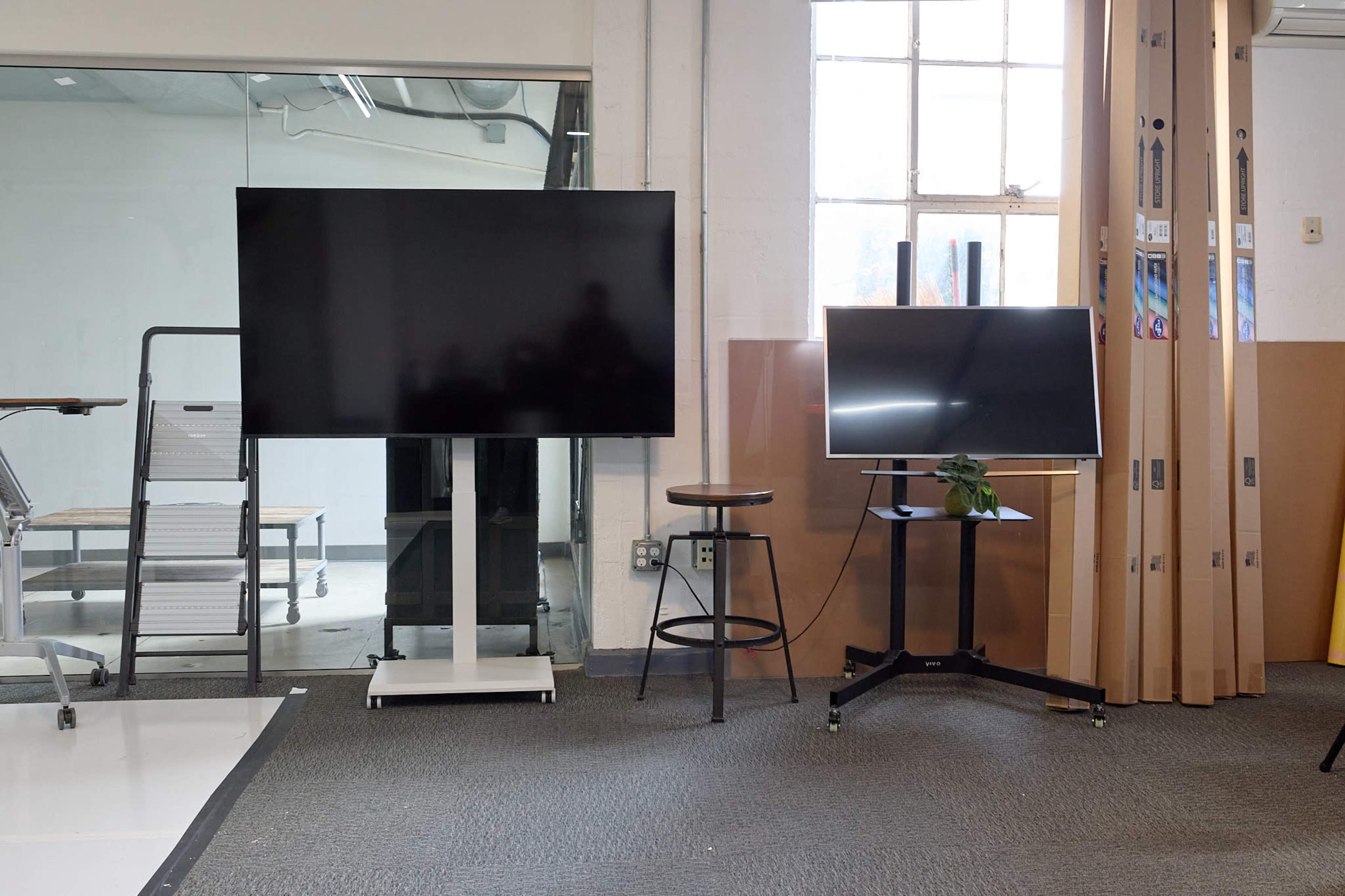 The image shows two television screens on stands, one large and black, positioned next to a smaller monitor, with a stool and a plant nearby, all set in a spacious room with exposed brick and large windows.