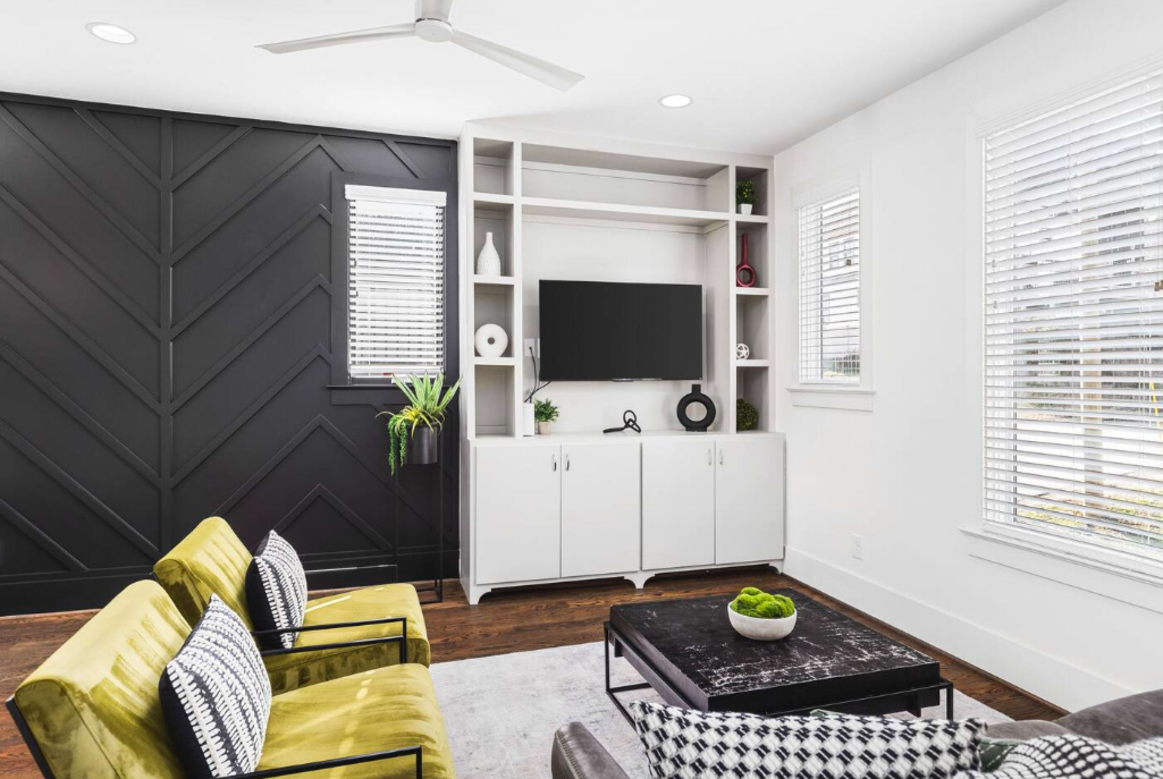 A modern living room featuring a black accent wall, a white television unit with shelving, a dark coffee table, and green and black upholstered chairs.