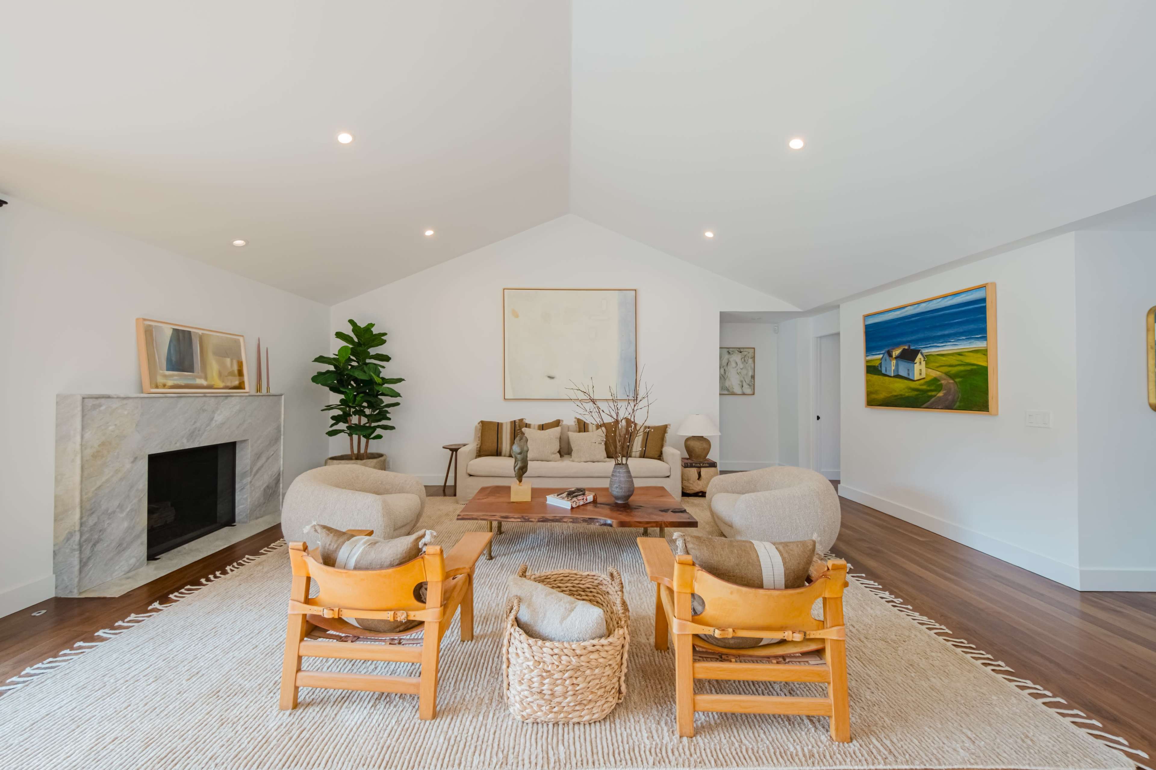 A bright living room features a marble fireplace, two armchairs, a sofa, a wooden coffee table, and artwork on the walls.