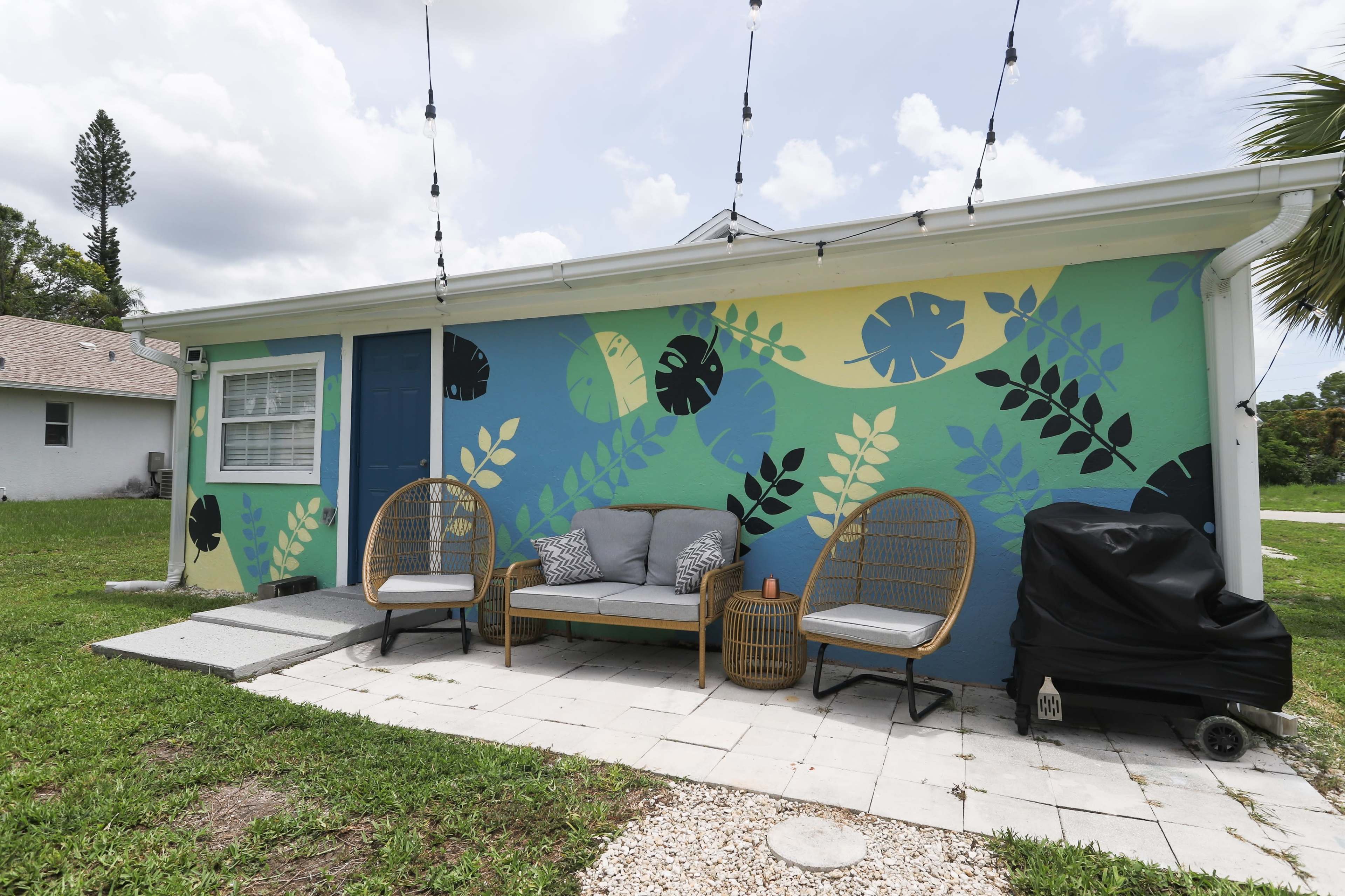 A colorful mural featuring plants and fish designs adorns the exterior wall of a house, with a seating area and a barbecue grill nearby.