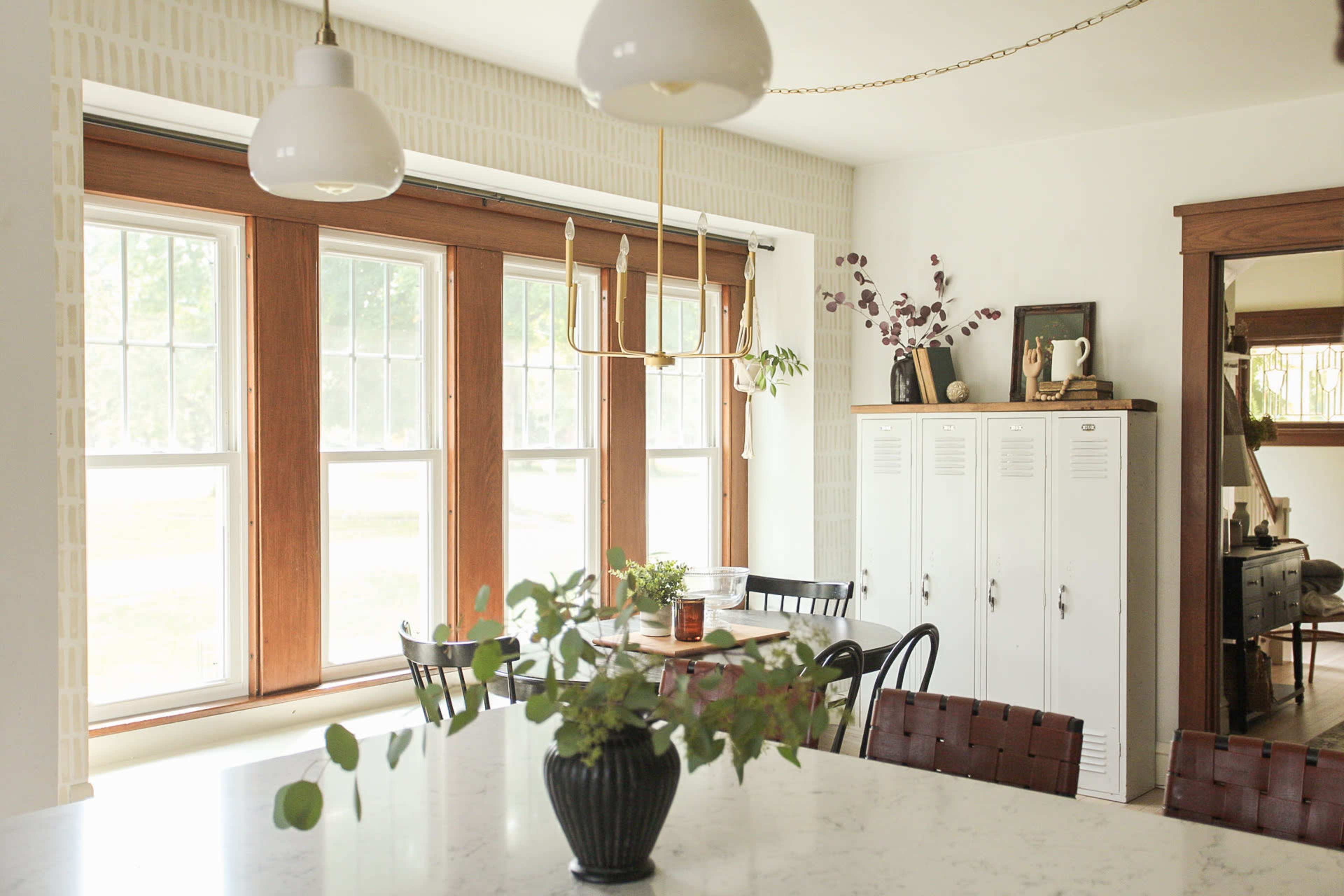 A bright kitchen features large windows, a dining table, black chairs, a white cabinet, and a vase with greenery on the counter.
