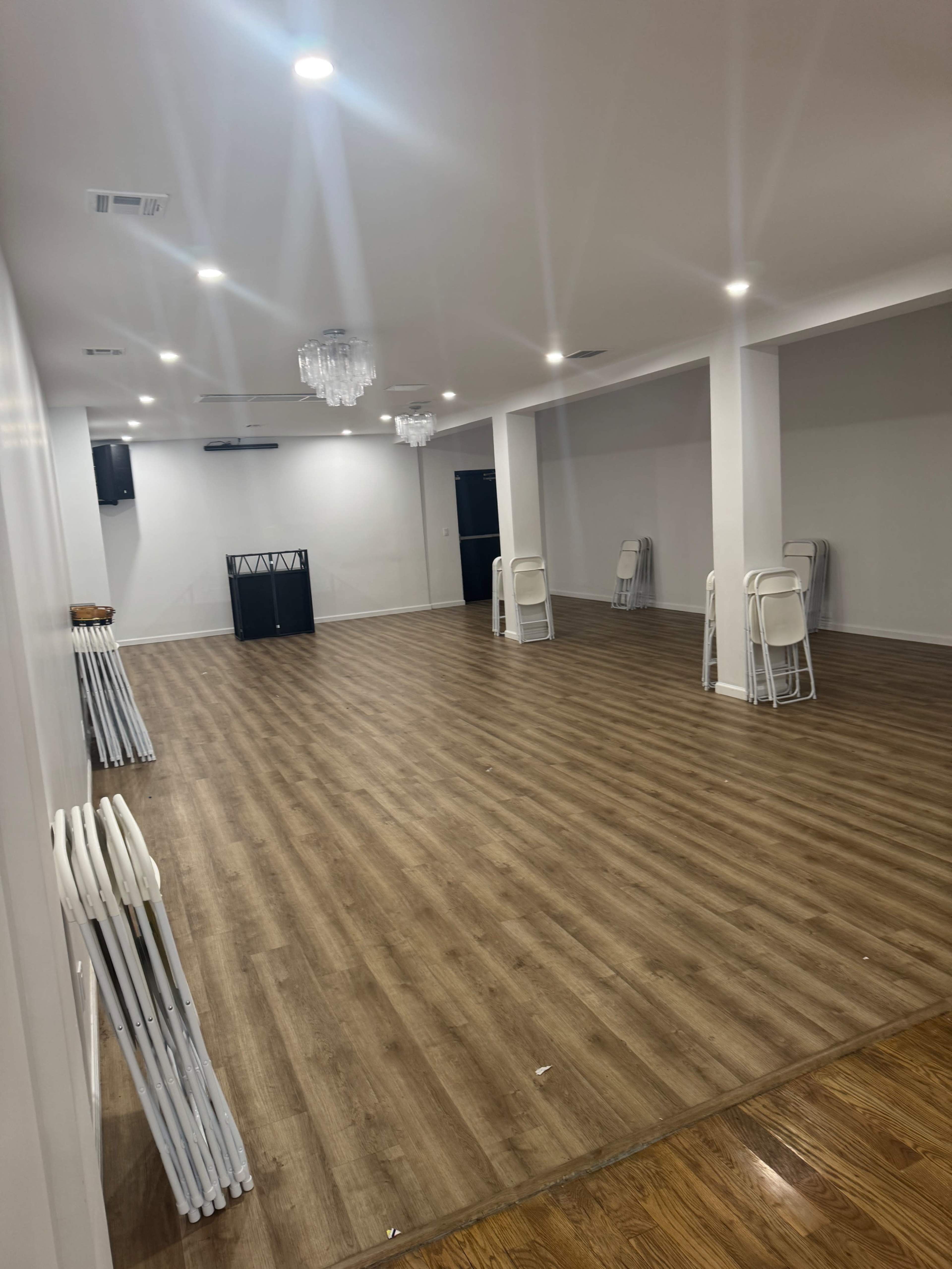 The image shows a large, empty interior space with wooden flooring, white walls, and several chairs stacked against one wall.