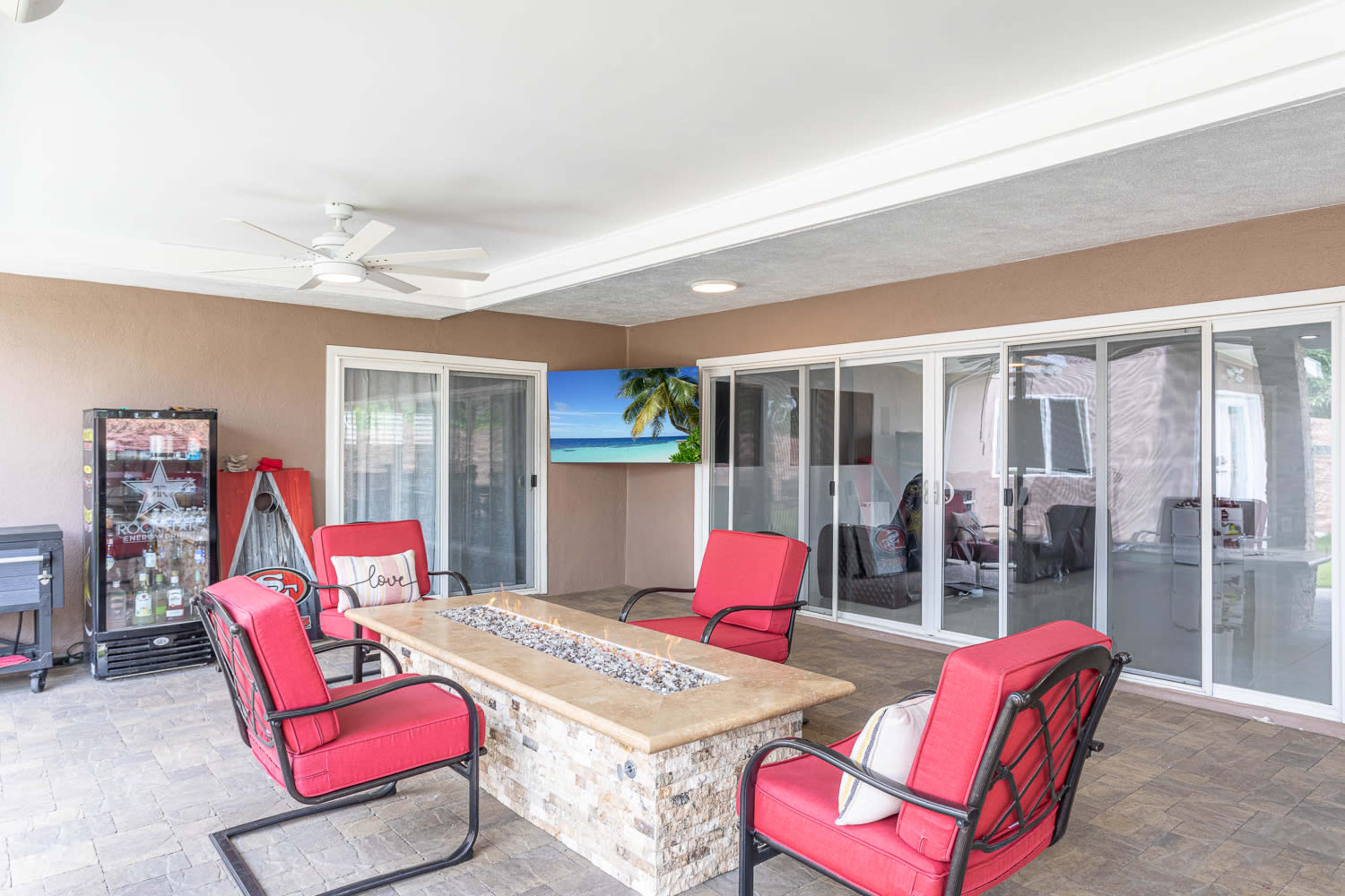 A covered patio with red chairs surrounding a stone fire pit, a sliding glass door leading to another room, and a tropical-themed artwork on the wall.