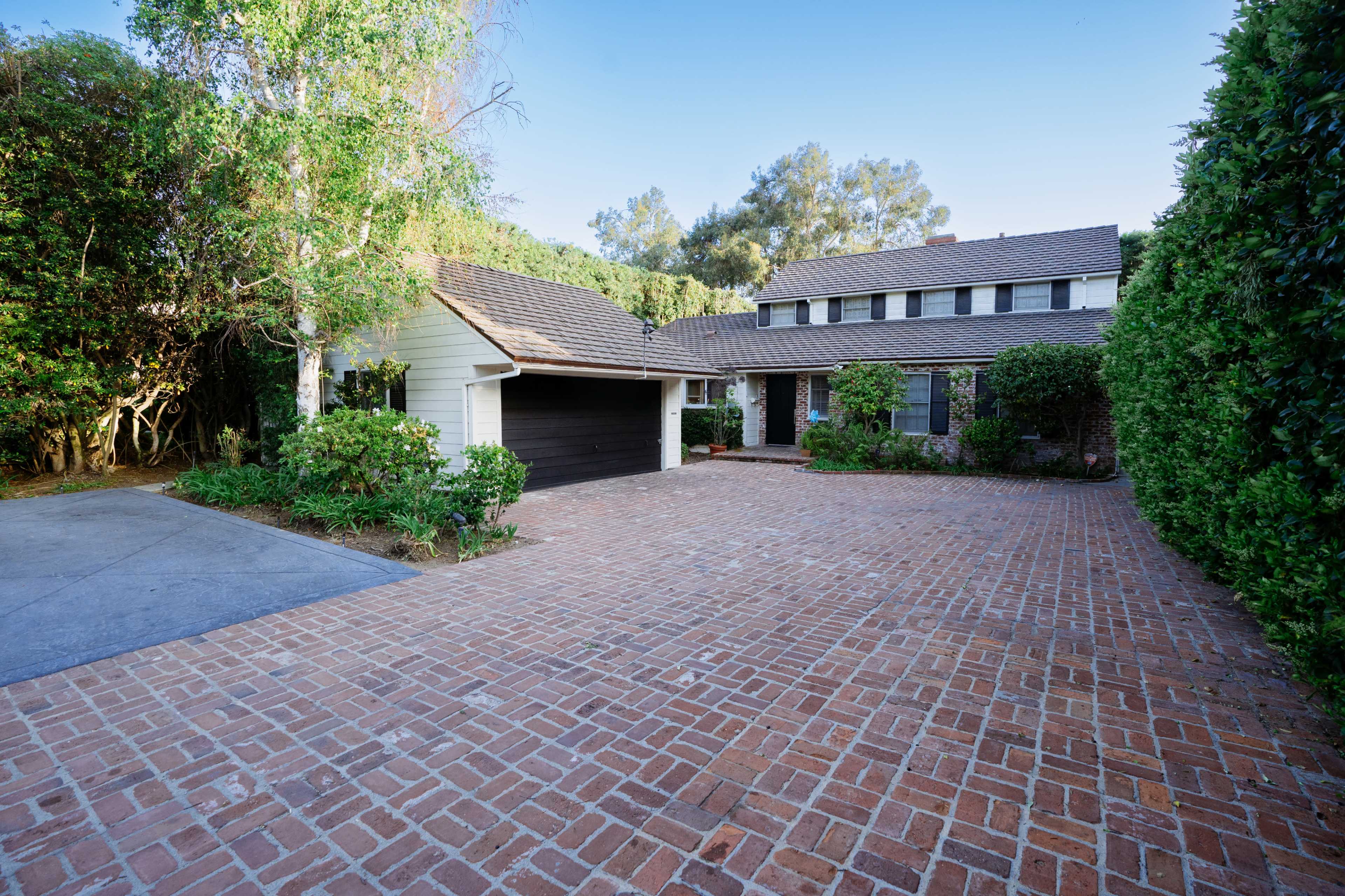 A two-story house with a brick driveway is surrounded by neatly trimmed hedges and trees.