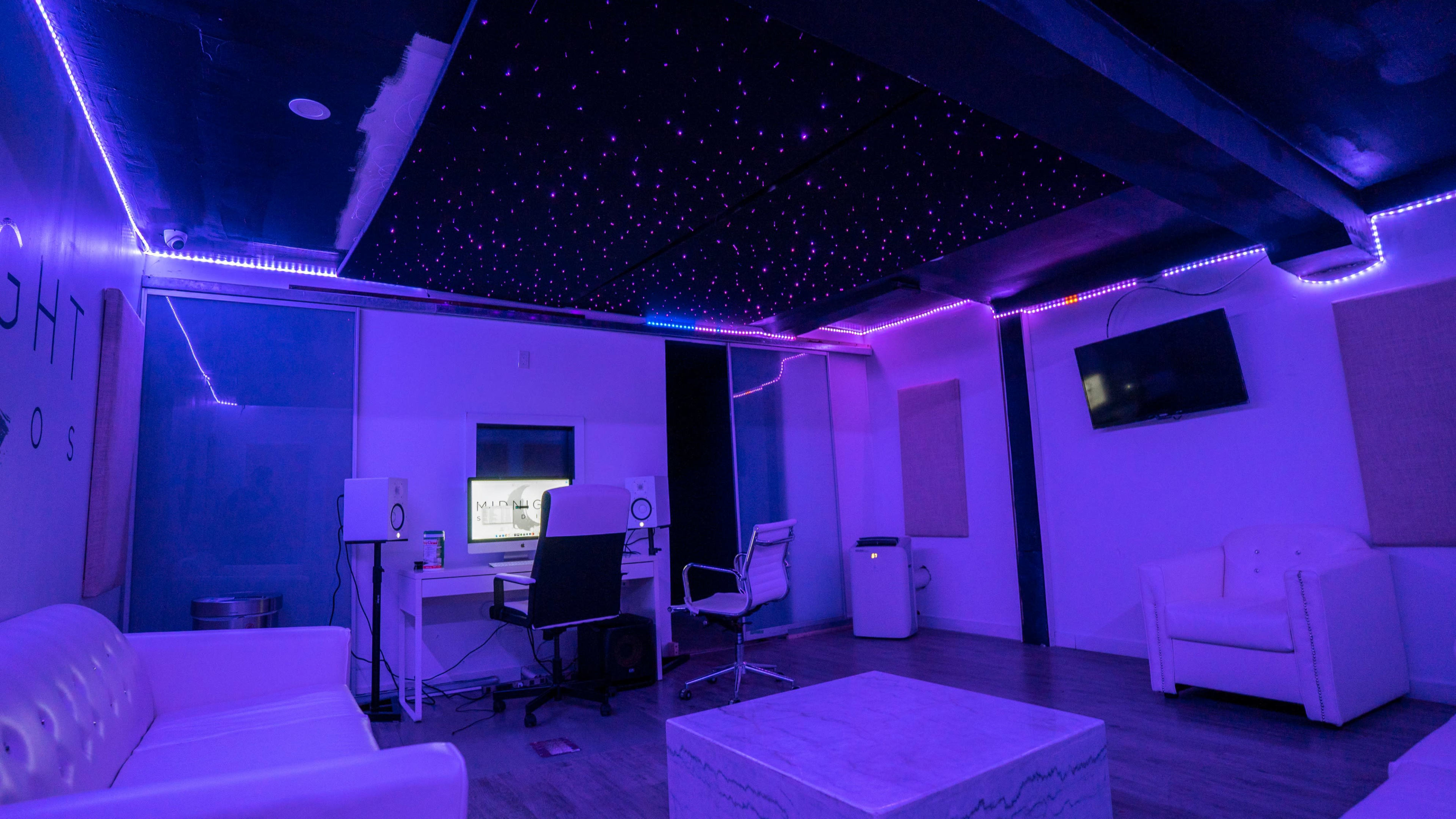A modern recording studio with purple lighting, a white sofa, a desk with studio monitors, and a screen in the background.
