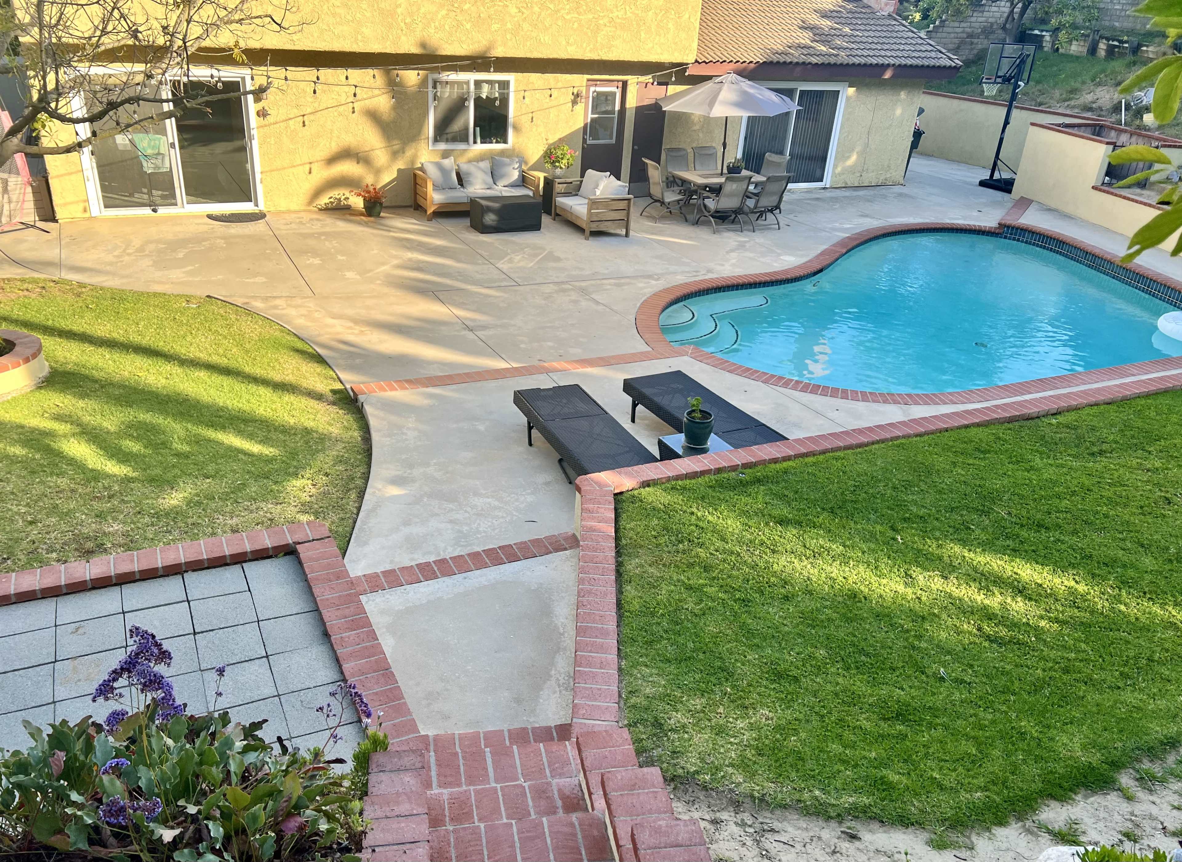 A poolside patio features a swimming pool, lounge chairs, and seating arrangements surrounded by landscaped grass and a stone pathway.