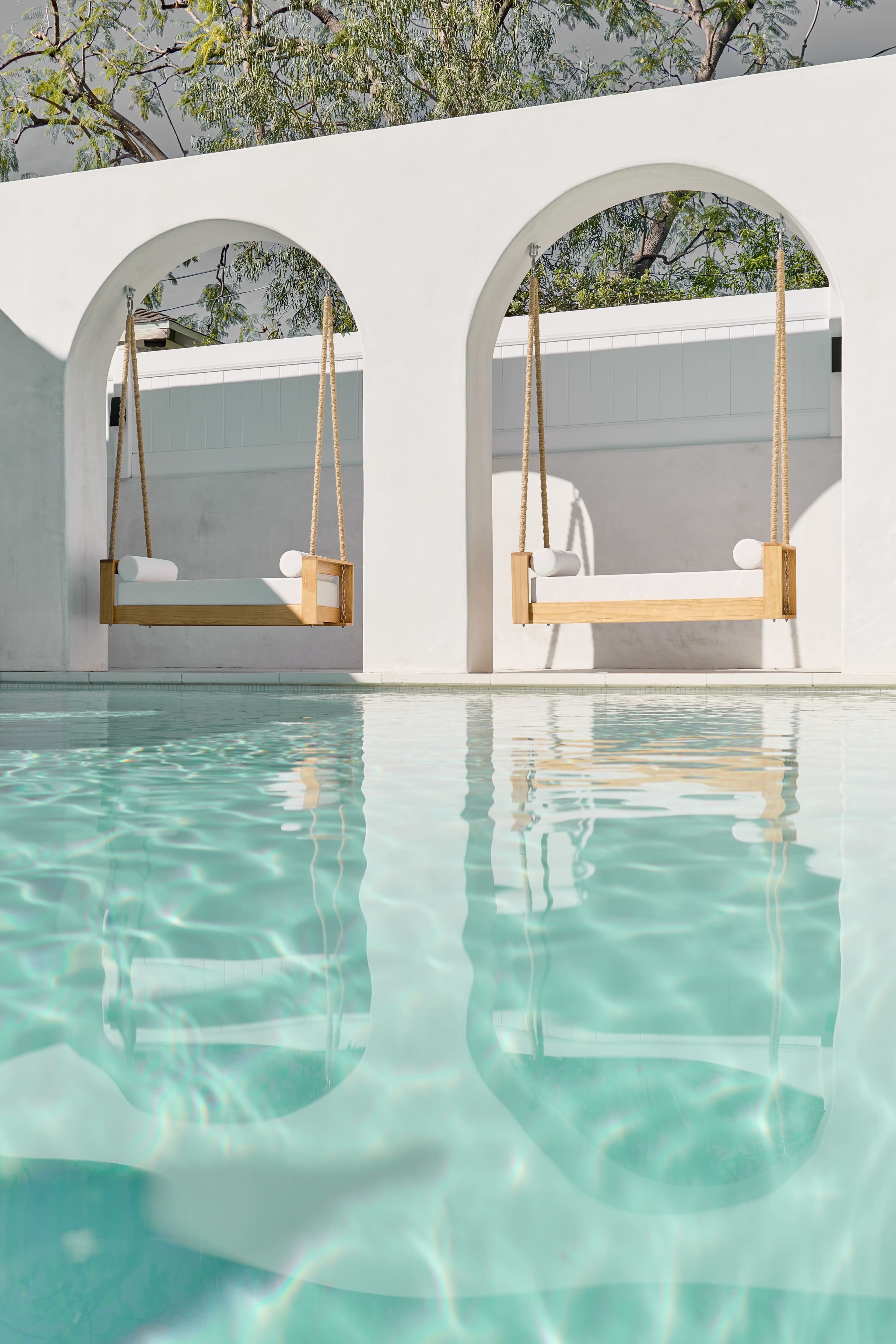 The image shows two wooden swings hanging in arched openings above a clear blue swimming pool, reflecting the surrounding architecture.