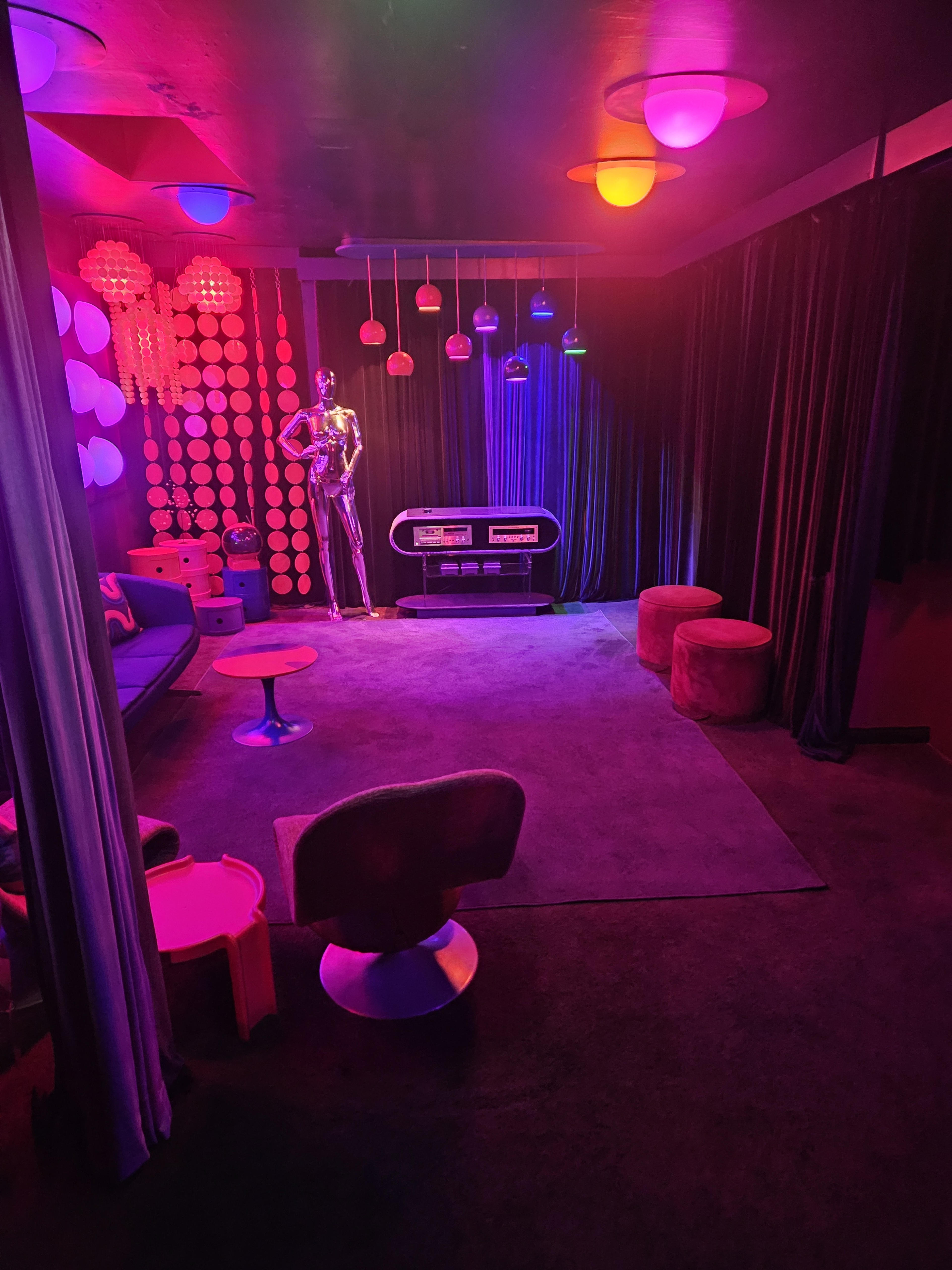 The scene features a vibrant, retro-themed room illuminated by colorful lighting, with plush seating, a mannequin, and decorative elements such as hanging light fixtures and wall decorations.
