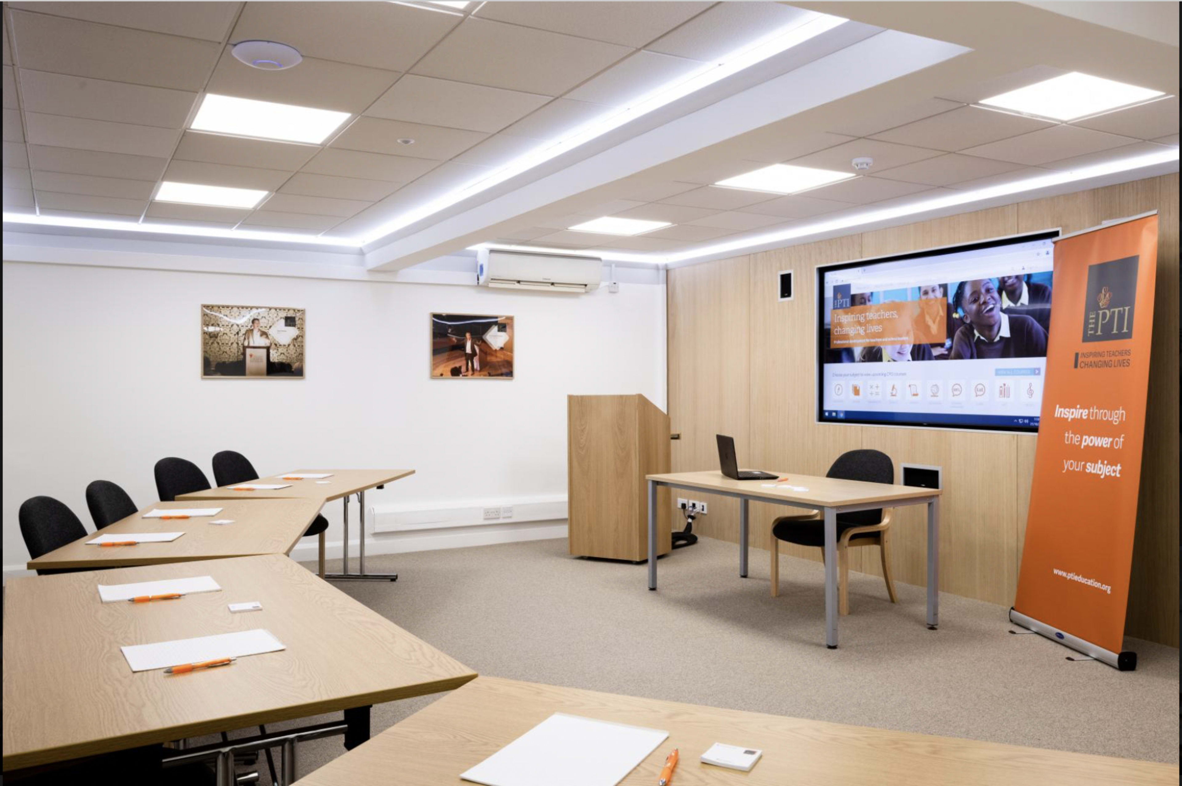 A modern training room features tables and chairs arranged for a meeting, with a large screen and a banner in the corner.