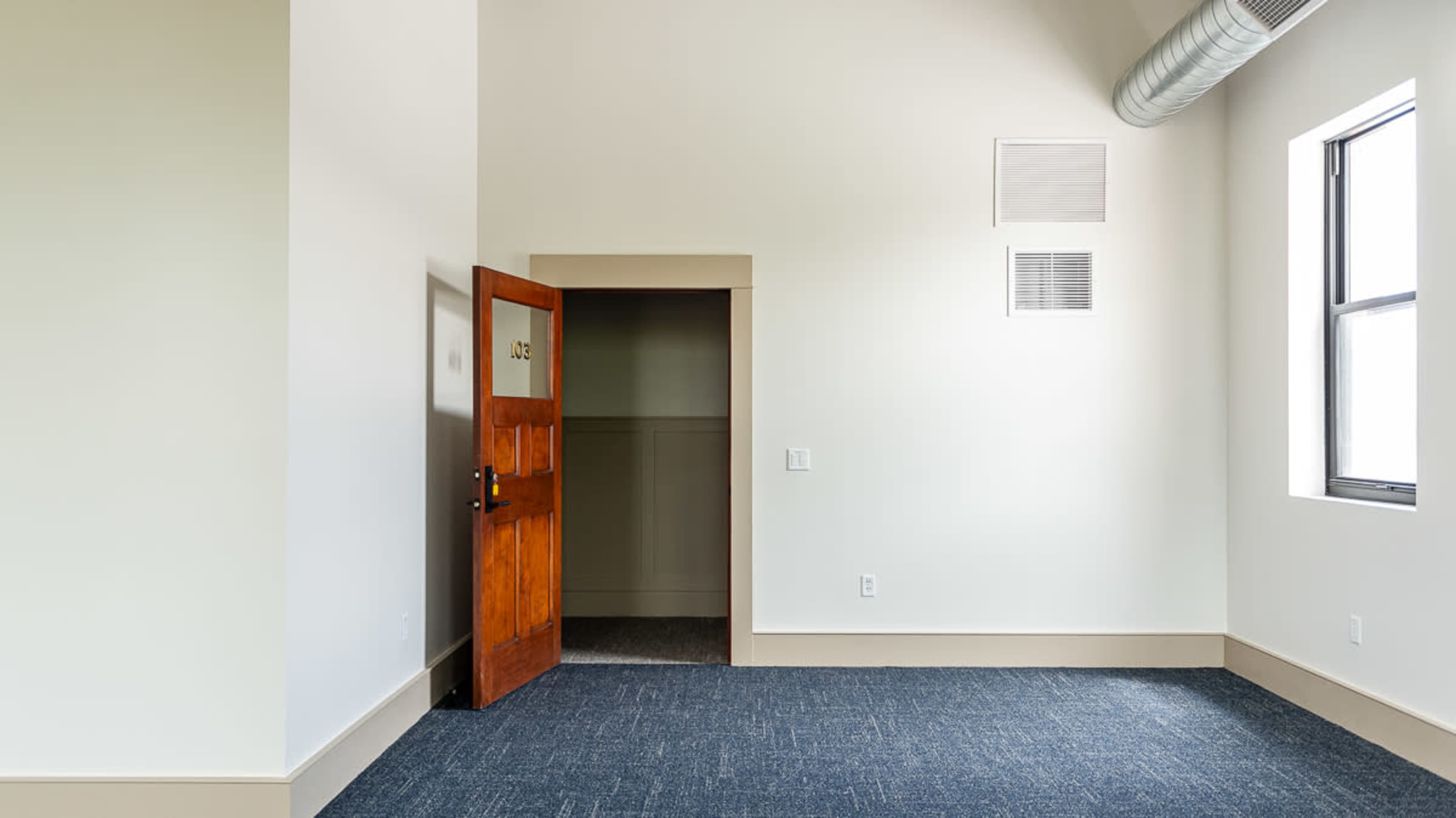 The image shows a simple room with a doorway leading to a small closet and a large window, featuring light-colored walls and carpeted flooring.