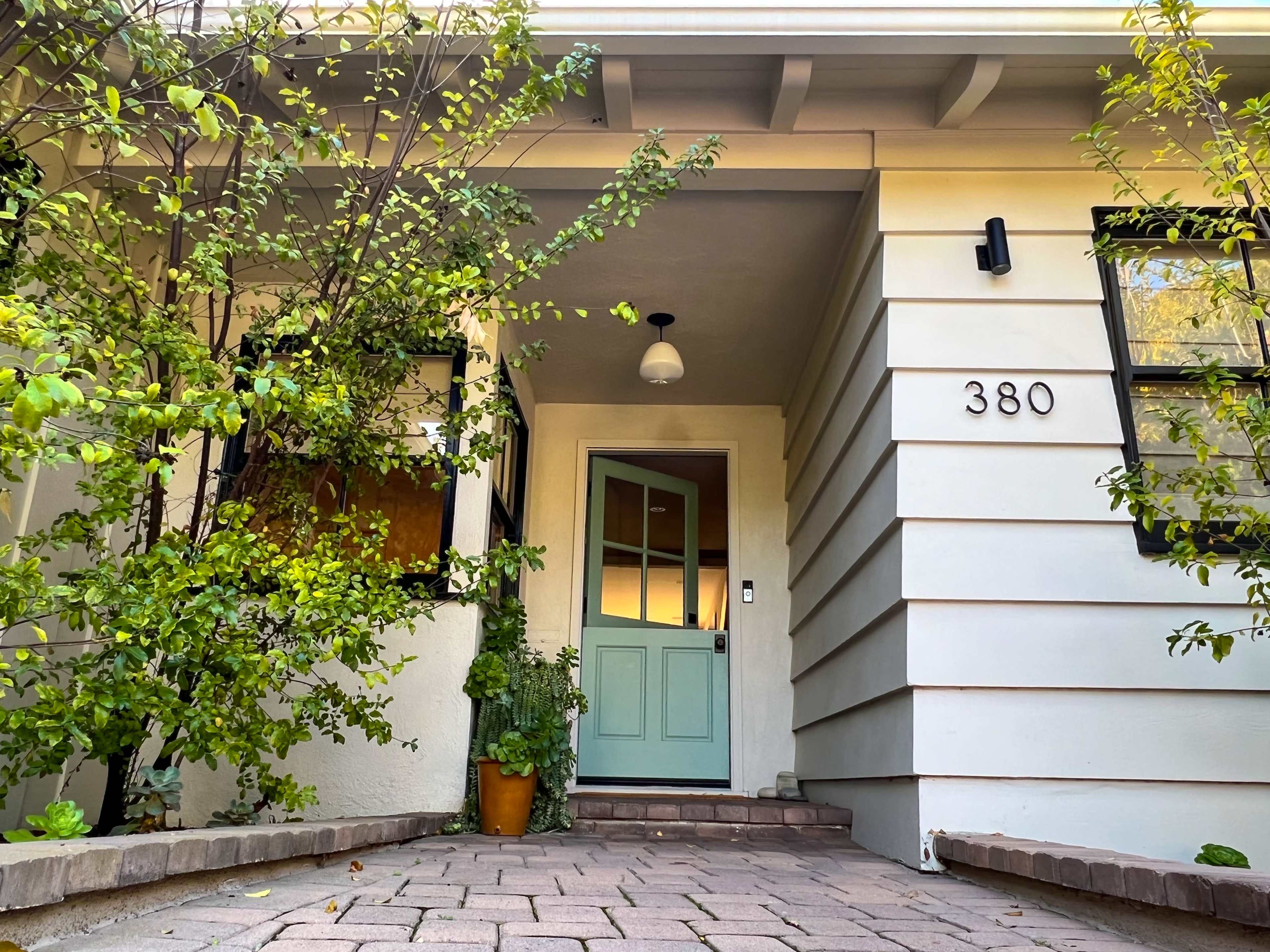 A house entrance with a light green door, surrounded by plants and a pathway made of pavers.